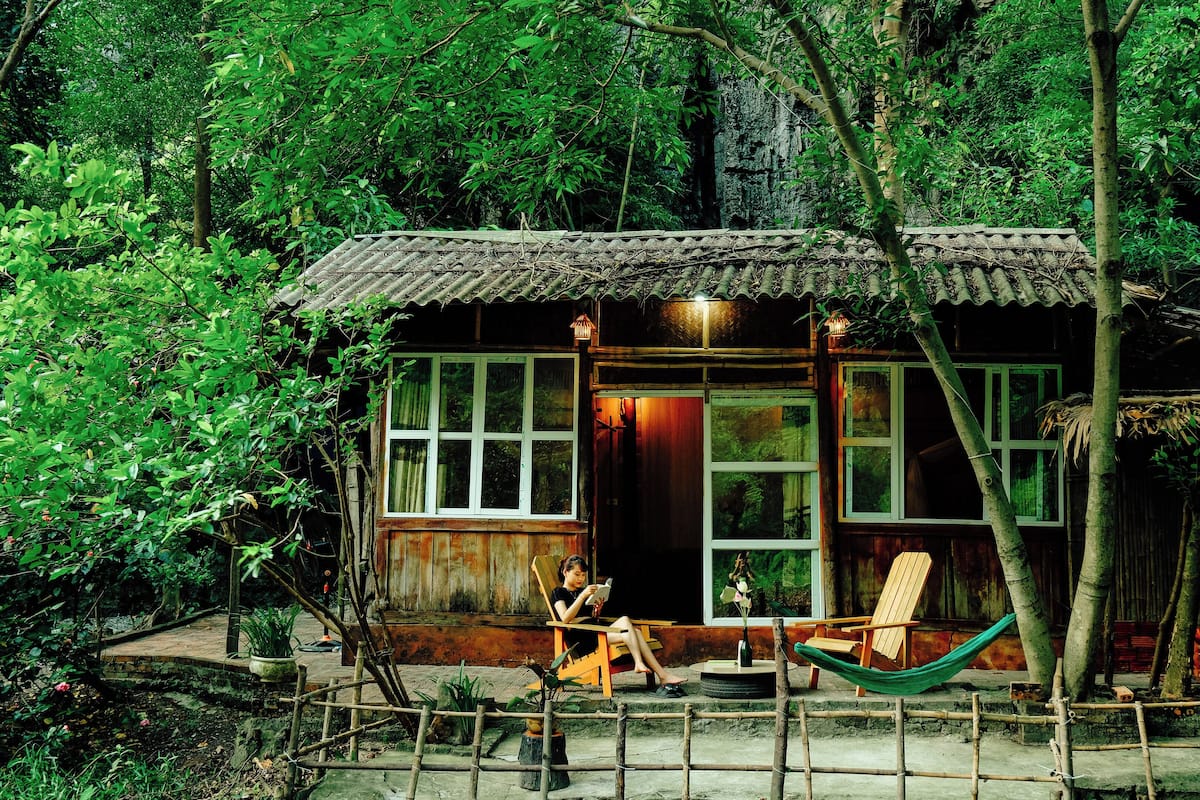 A rustic wooden cabin with large windows, a hammock, and a small outdoor seating area surrounded by lush greenery.