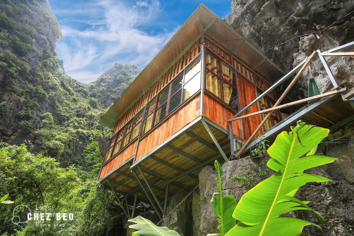 A wooden structure with a balcony, surrounded by lush greenery and rocky cliffs.