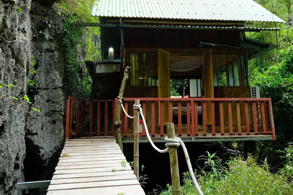A wooden bridge leading to a wooden cabin with a metal roof, surrounded by lush greenery and rocky cliffs.