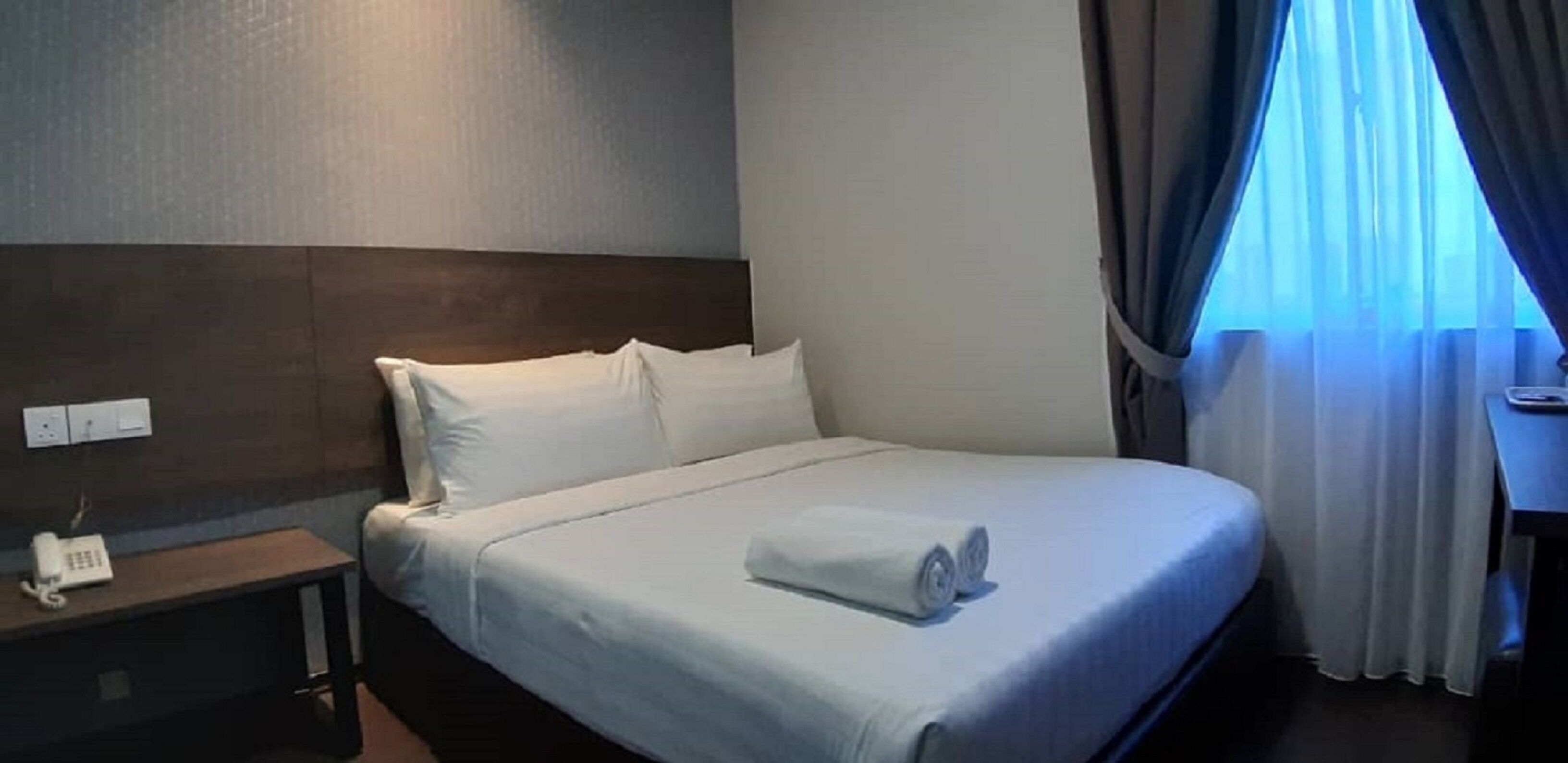 Ett hotellrum med en väl bäddad säng, ett nattduksbord med en telefon och ett fönster med gardiner.