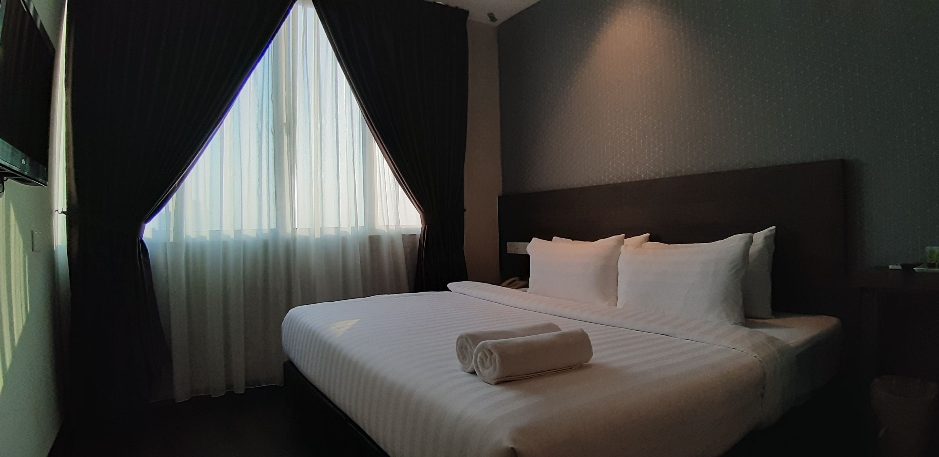 Une chambre d’hôtel avec un grand lit, du linge de lit blanc, une tête de lit et une fenêtre ornée de rideaux légers.