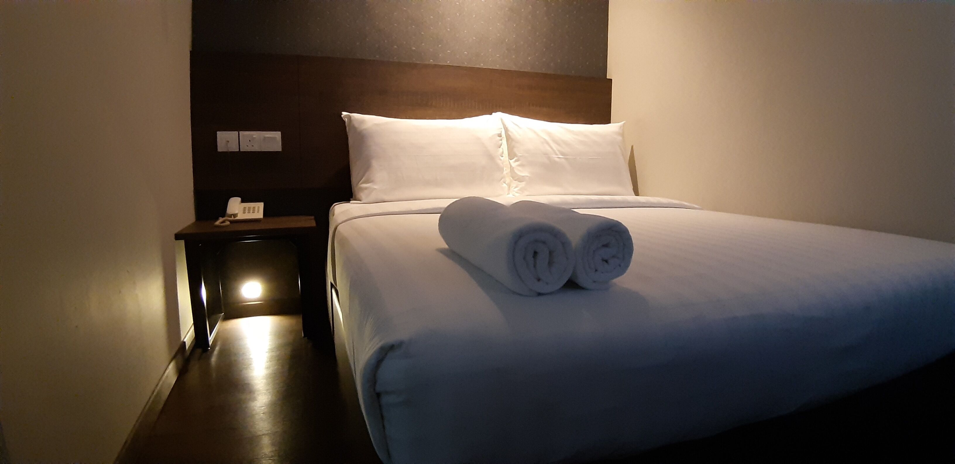 A hotel room with a neatly made bed, a bedside table with a lamp, and a wall-mounted power outlet.