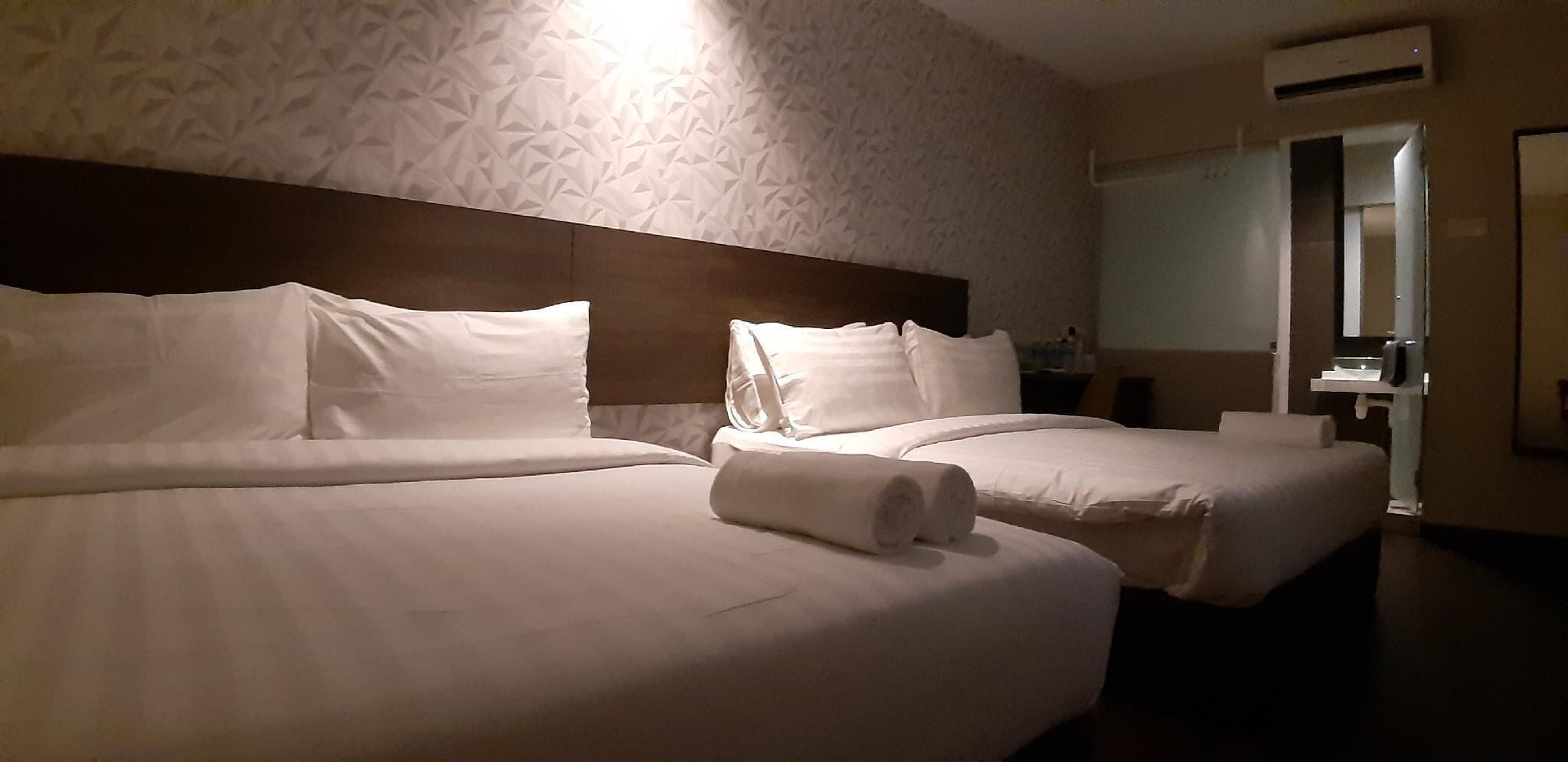 Hotellihuone, jossa on kaksi sänkyä, sängynpääty, peili, pesuallas ja ilmastointilaite.