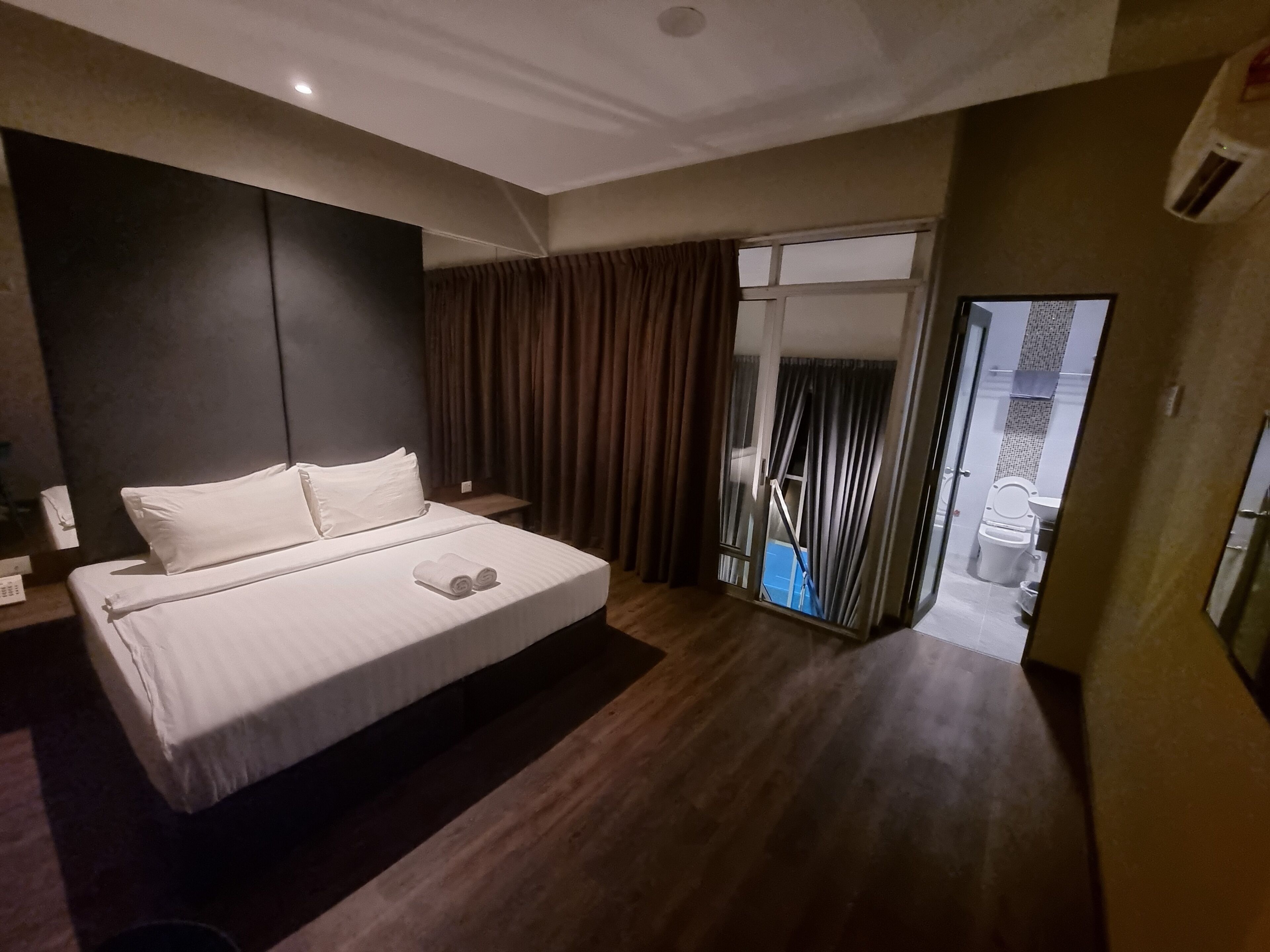 A modern hotel room with a large bed, a small desk, a bathroom visible through a glass door, and a wooden floor.