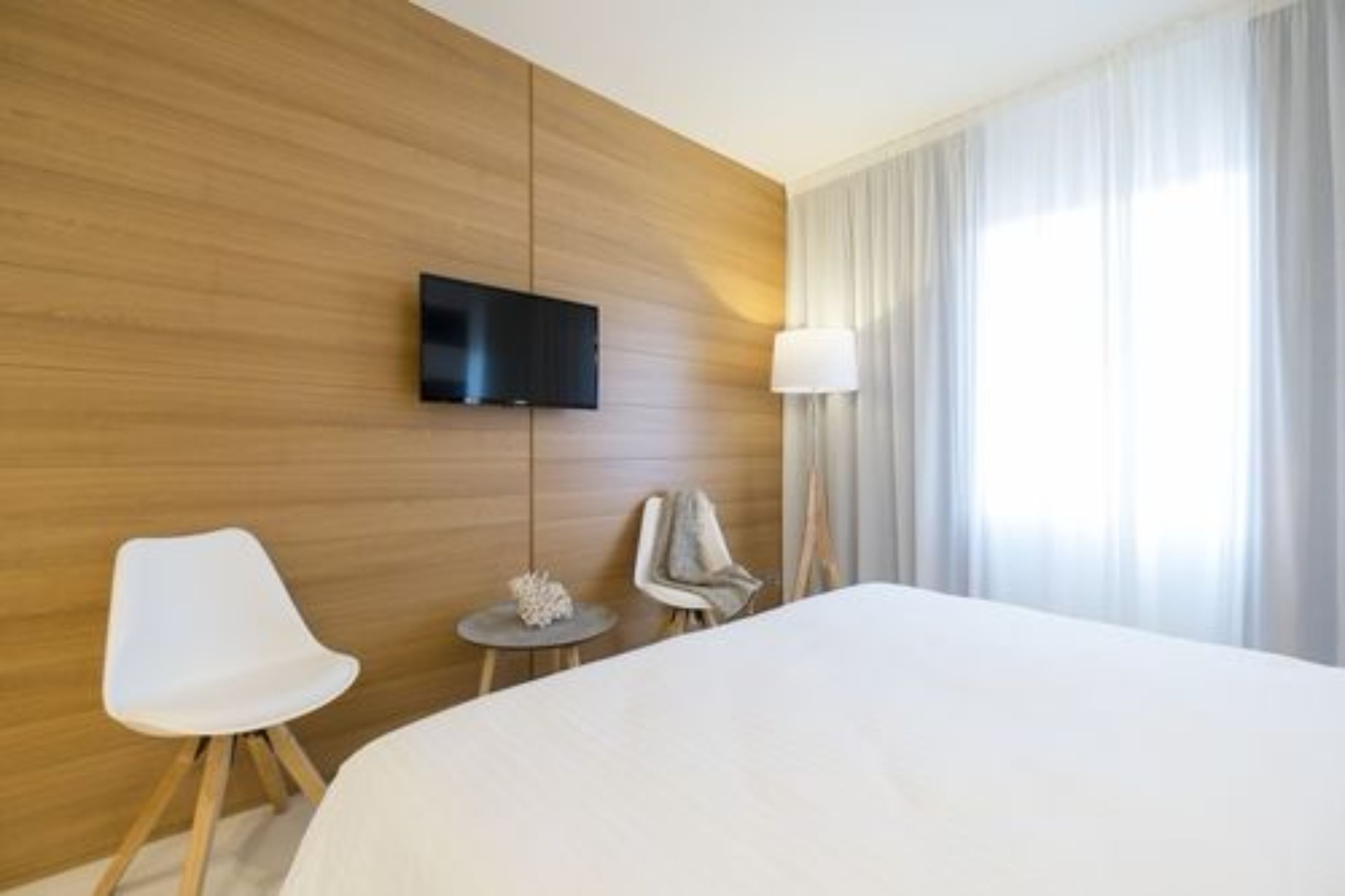Een hotelkamer met een groot bed, een flatscreen televisie aan de muur, een klein rond tafeltje met een lamp en een witte stoel.