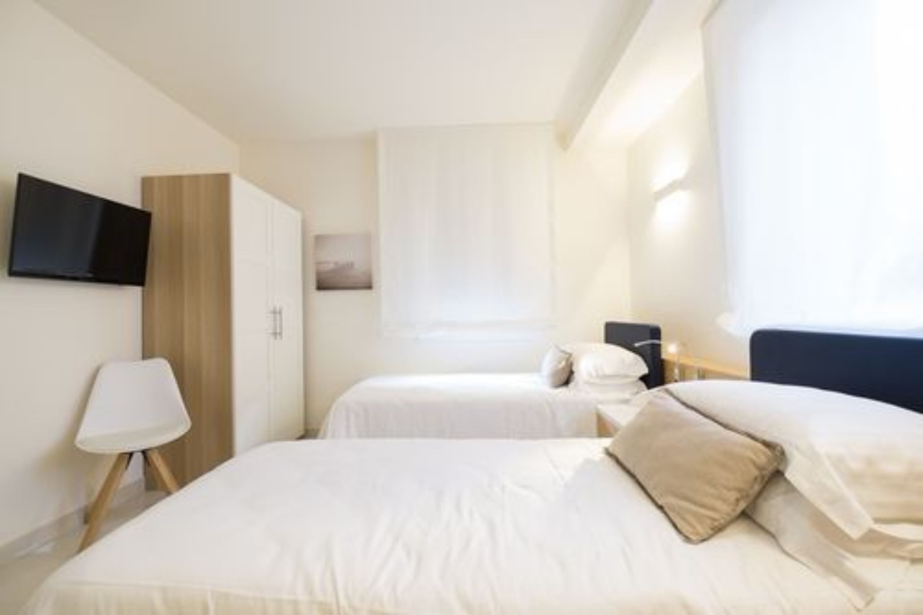 Camera d'albergo con due letti, una TV a muro, un comodino con una lampada e una finestra con tende.