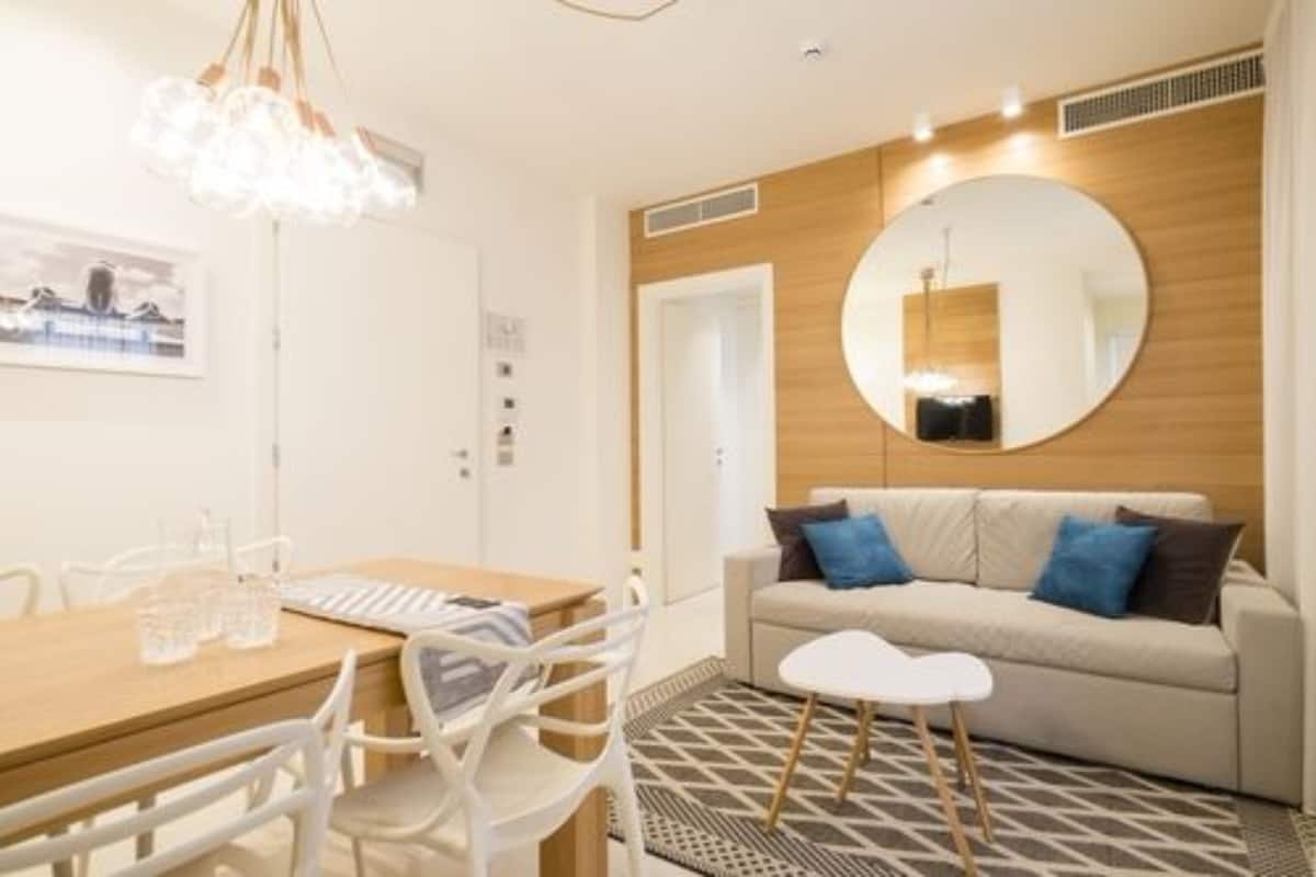 A modern room with a wooden accent wall, a round mirror, a sofa with blue cushions, a white coffee table, and a dining area with a wooden table and chairs.