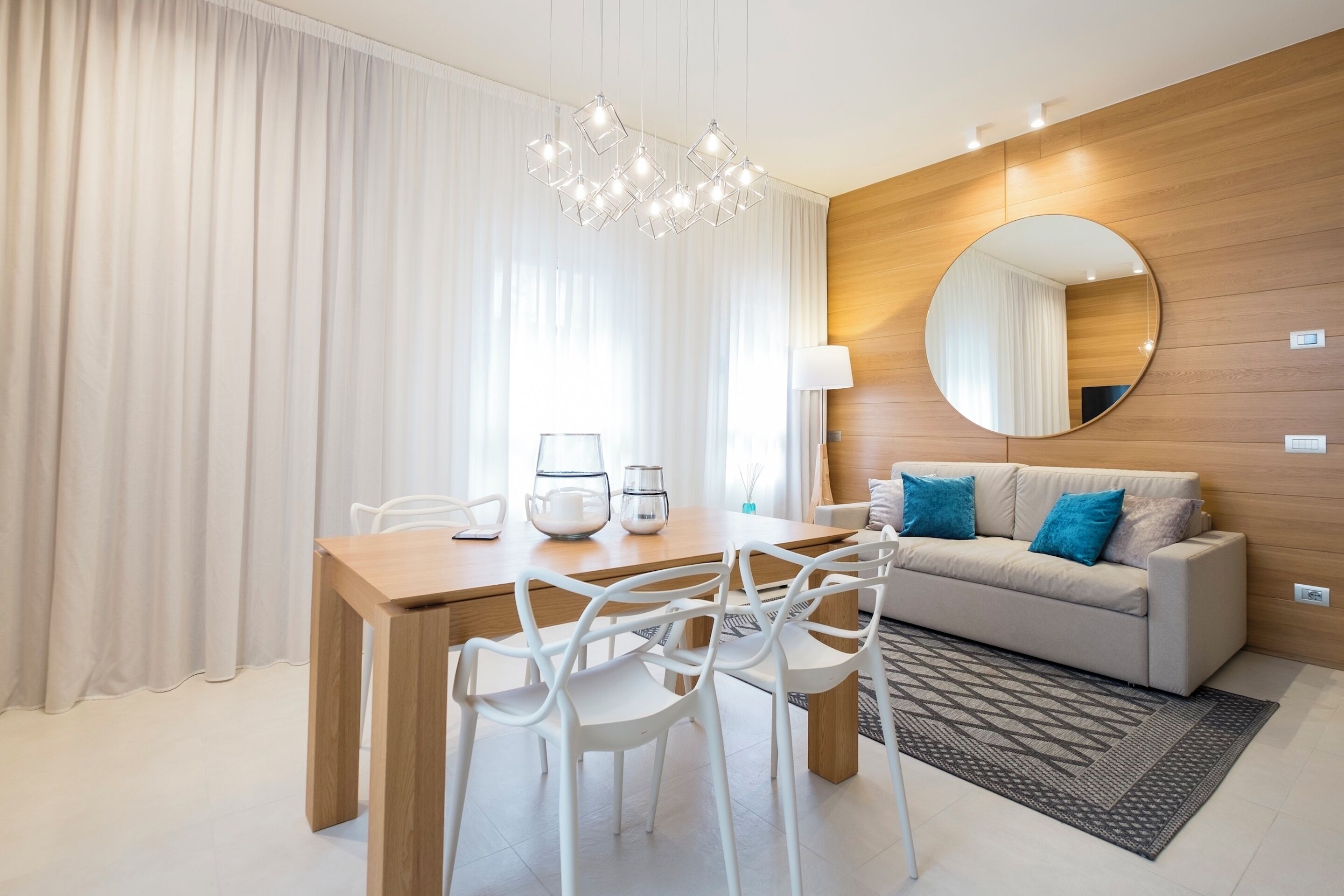 A modern hotel room with a wooden accent wall, a round mirror, a dining table with chairs, and a sofa with blue cushions.