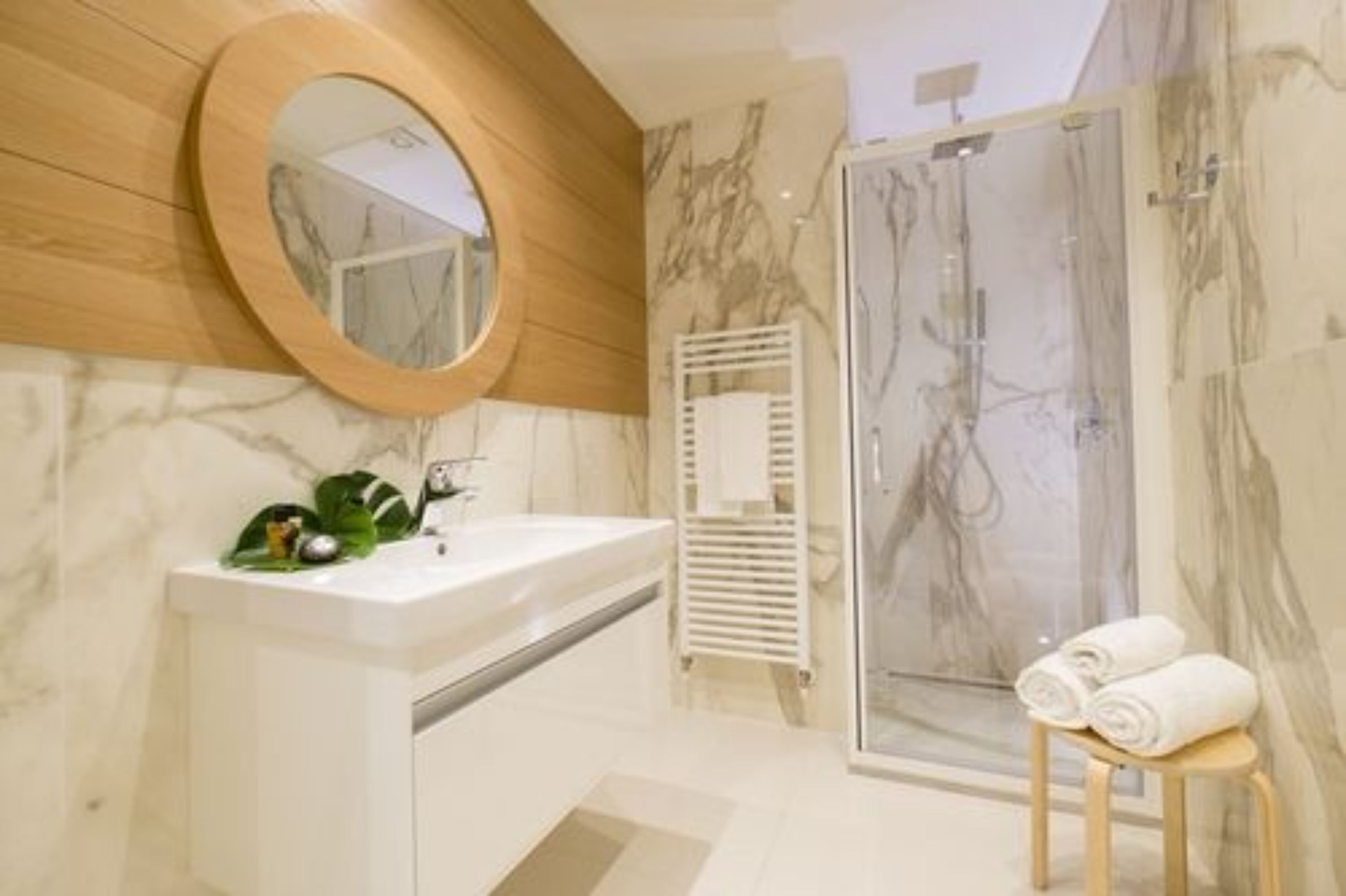 A modern bathroom with a marble vanity, a wooden-framed mirror, a glass shower enclosure, and a towel stand.