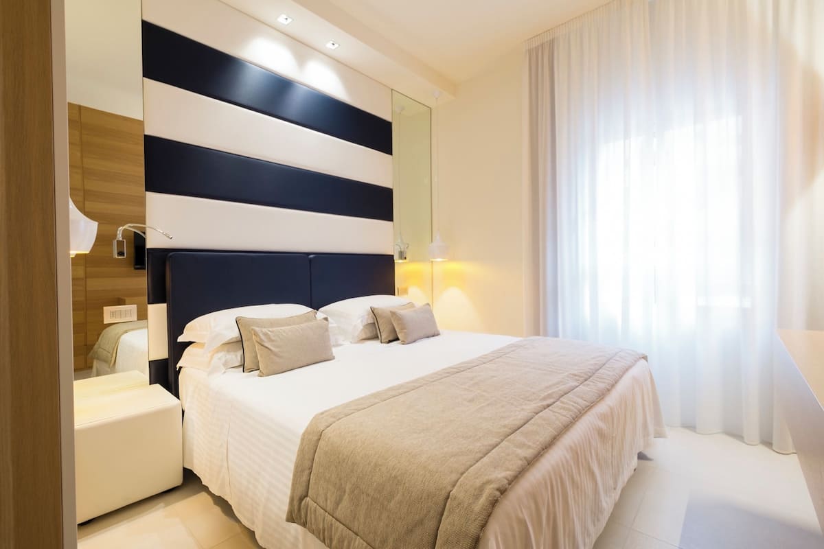 A modern hotel room with a large bed, a bedside table, a mirror, and a window with curtains.
