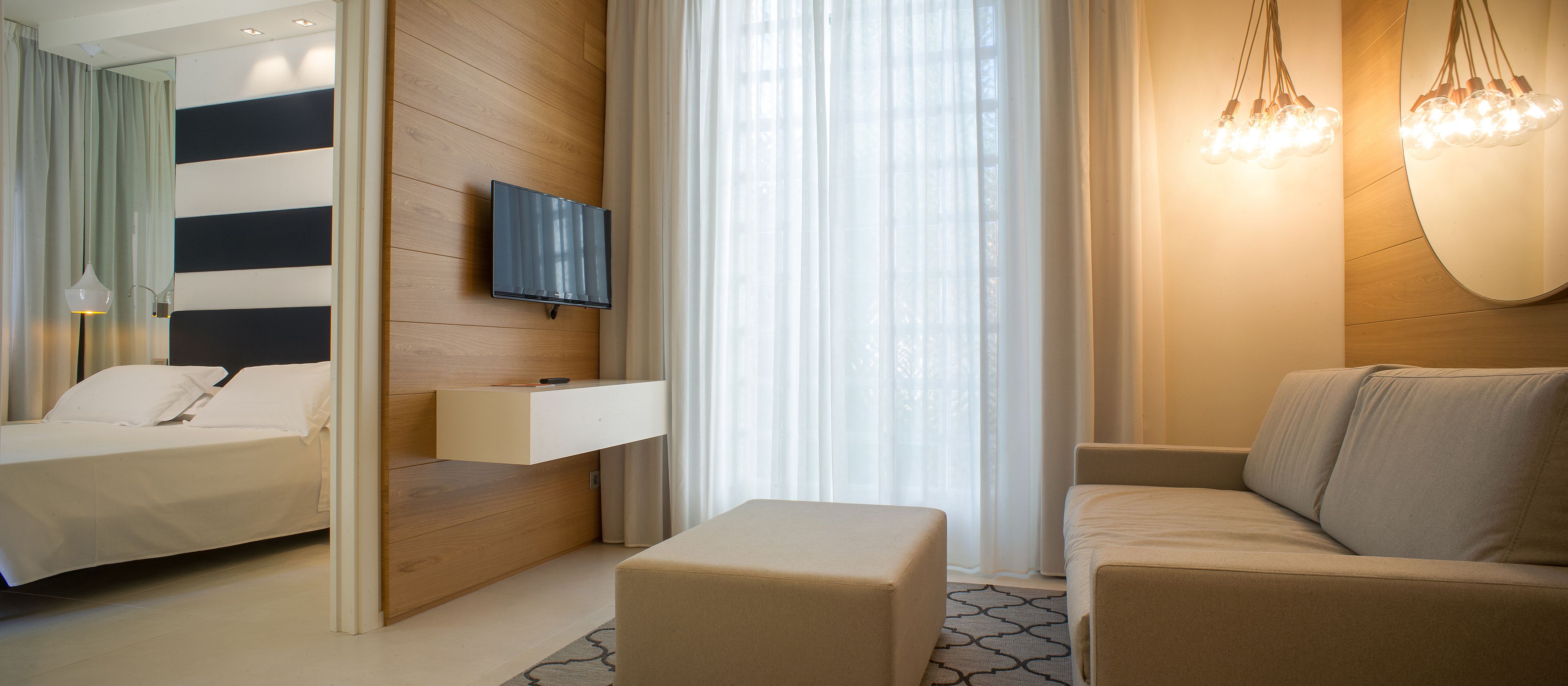 A modern hotel room with a bed, a sofa, a TV mounted on the wall, and a hanging light fixture.