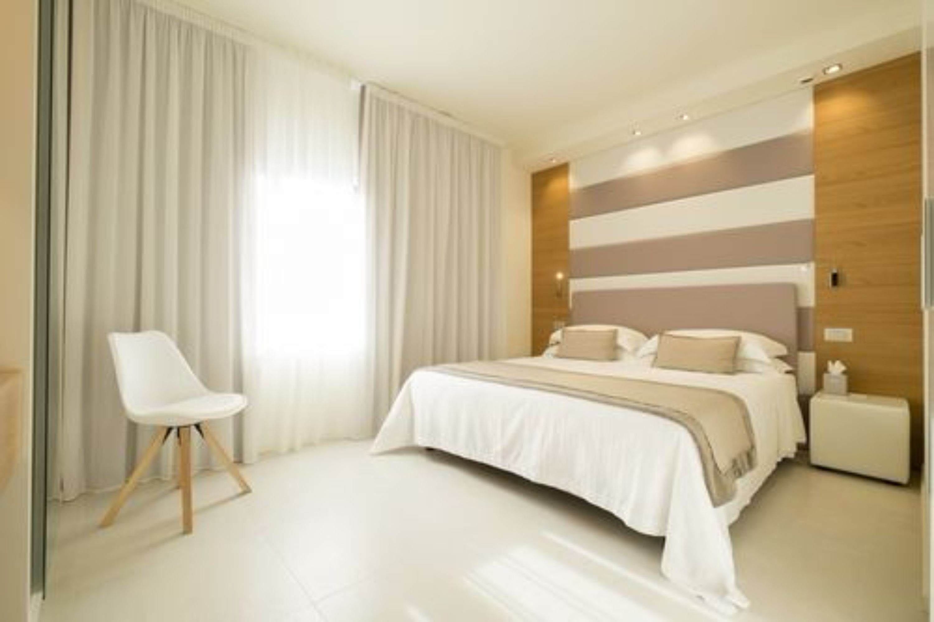 Une chambre à coucher moderne avec un grand lit, une chaise blanche et une vue sur l’extérieur à travers des rideaux transparents.