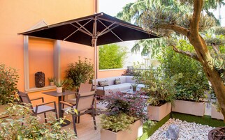 Un patio con tavolo, sedie e ombrellone. Sono presenti piante in vaso e un piccolo giardino con rocce e un albero.
