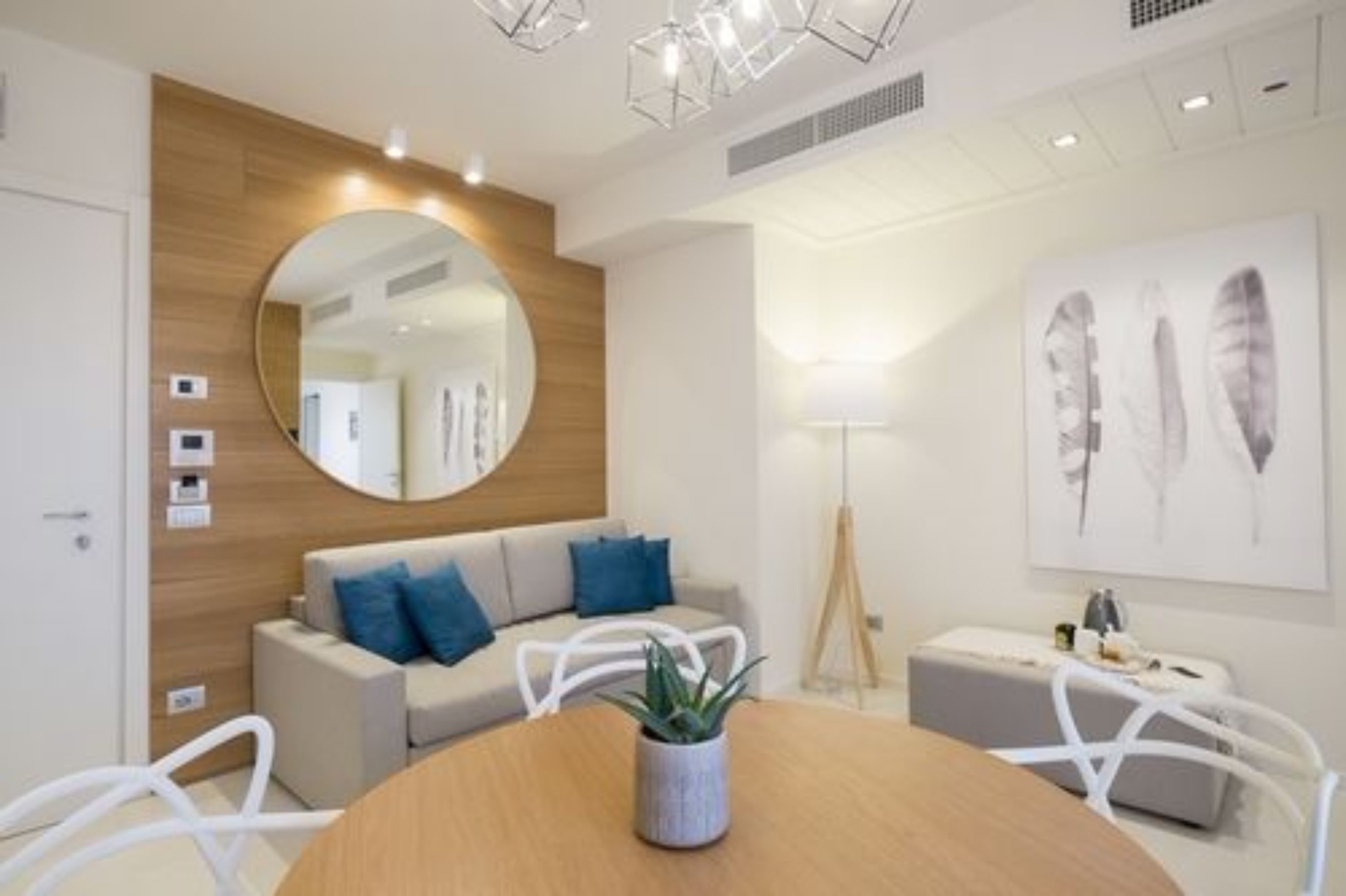 Una sala moderna con una mesa redonda de madera, un sofá con cojines azules, un espejo redondo y una lámpara de pie.