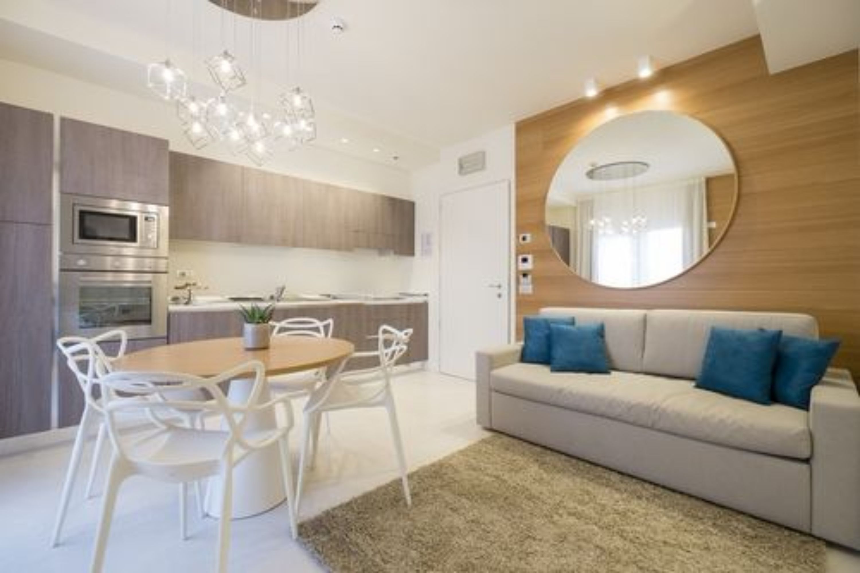 A modern kitchen with built-in appliances, a dining area with a round table and chairs, and a living area with a sofa and a round mirror.
