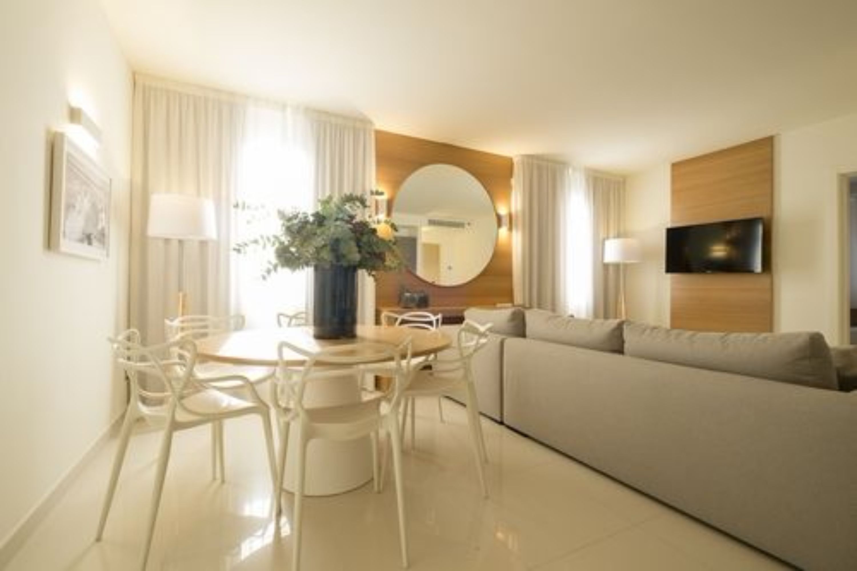 A modern hotel room with a dining area, a sofa, a TV mounted on the wall, and a large mirror.