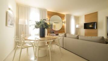 A modern hotel room with a dining area, a sofa, a TV mounted on the wall, and a large mirror.