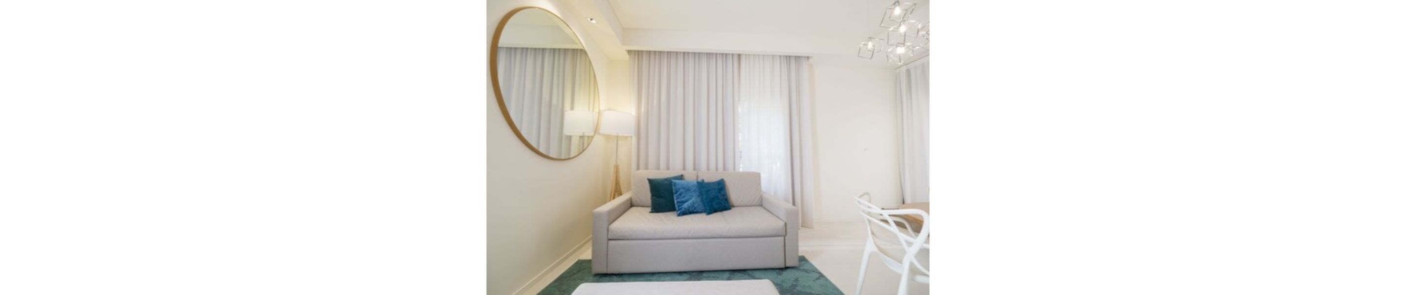 A modern hotel room with a sofa, a round mirror, a chandelier, and a view of the outside through large windows.