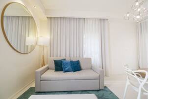 A modern hotel room with a sofa, a round mirror, a chandelier, and a view of the outside through large windows.
