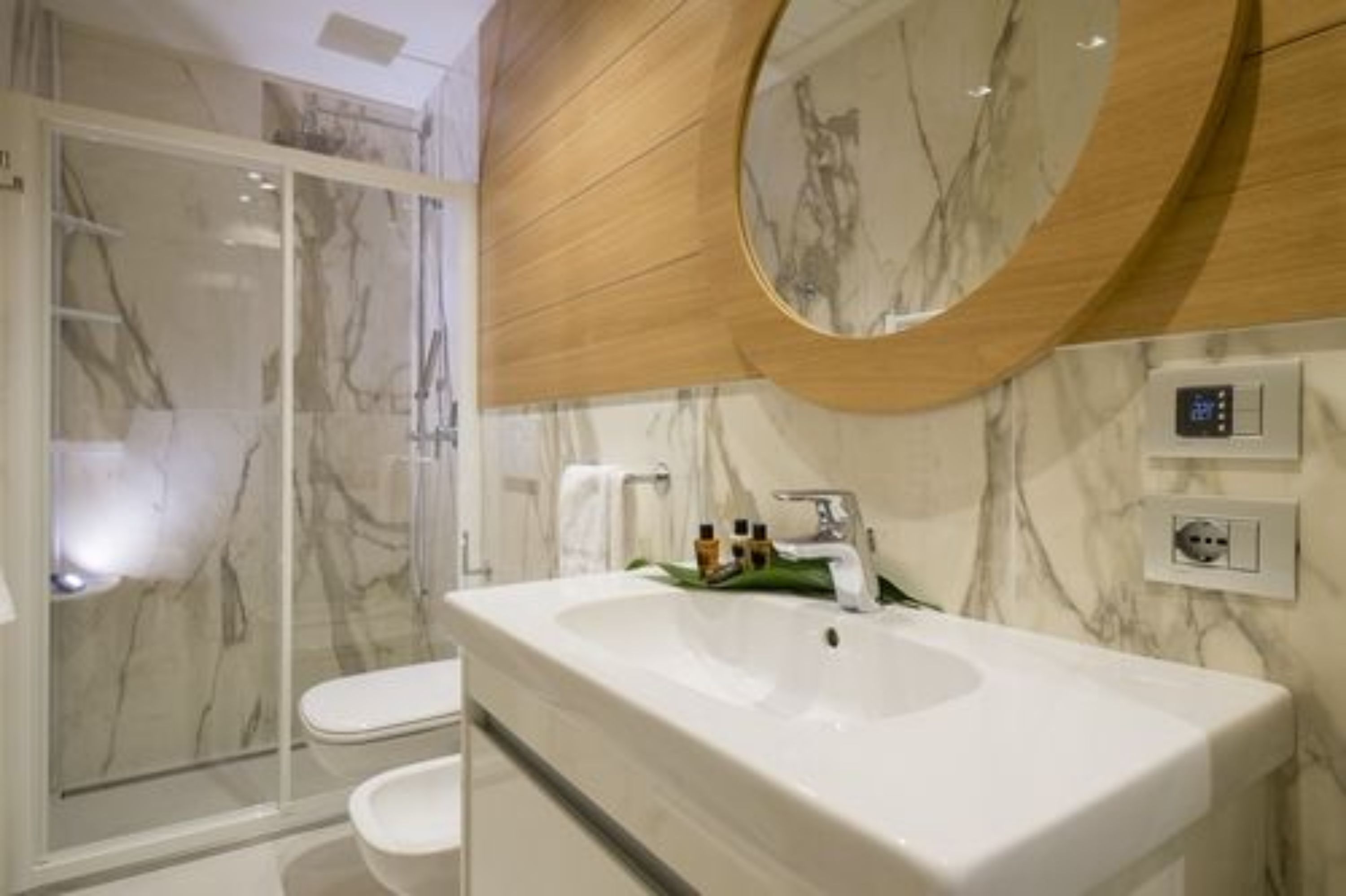 Ein modernes Badezimmer mit Marmorwaschbecken, einem Holztischchen, einer Glasdusche und einem runden Spiegel.