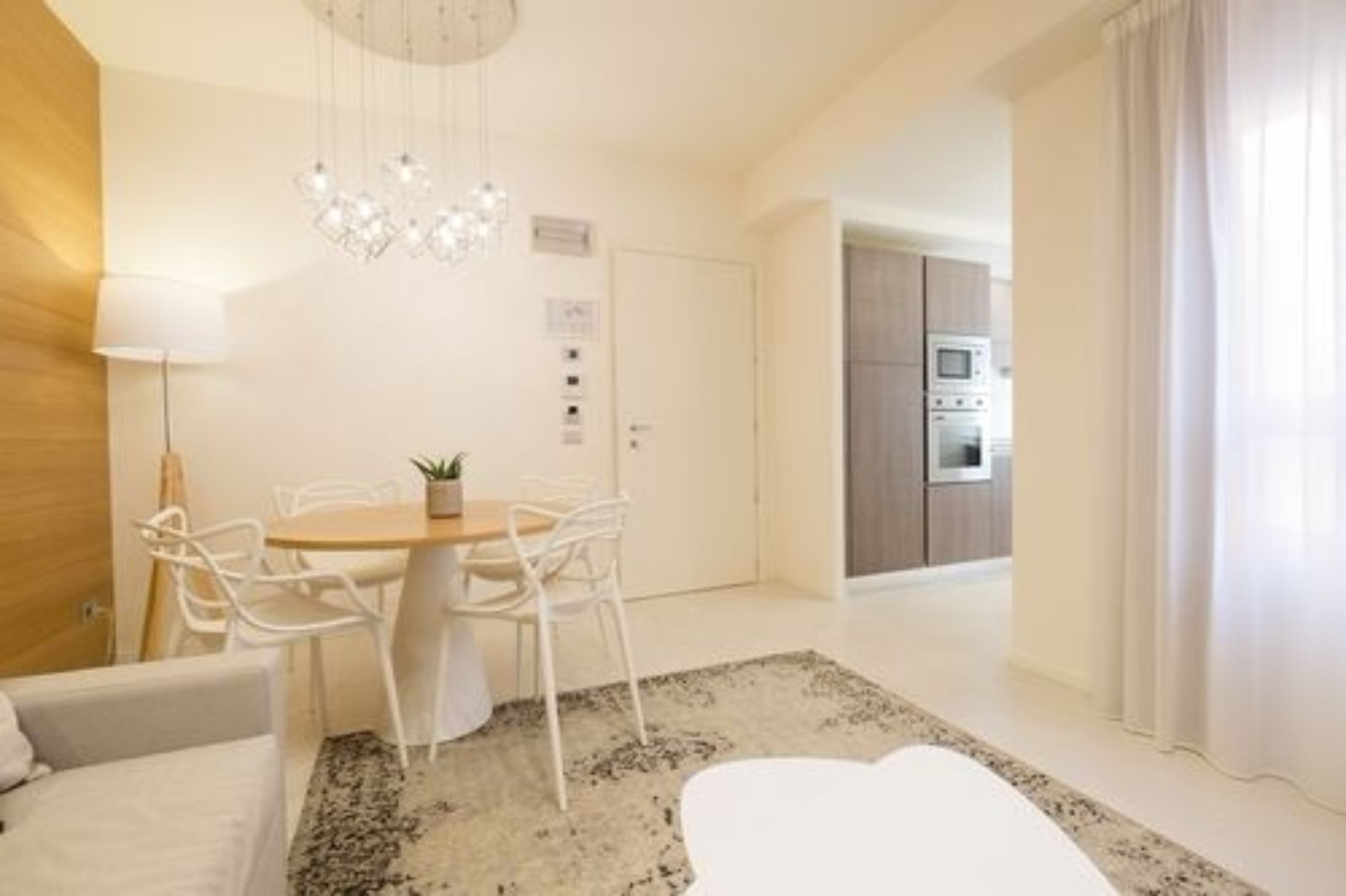 Un salon moderne avec une table de salle à manger ronde, des chaises blanches, un lustre et une vue sur la cuisine.