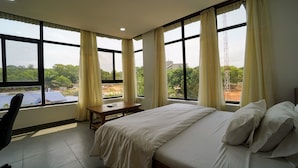 Habitación de hotel con una cama grande, un escritorio y una silla. Varias ventanas con cortinas ofrecen vistas a zonas verdes y edificios.