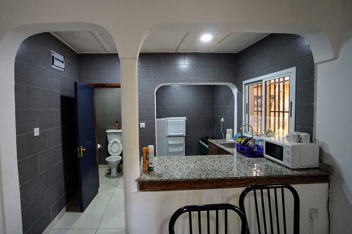 A compact bathroom with dark tiles, a white toilet, a sink with a mirror, and a microwave on the countertop.
