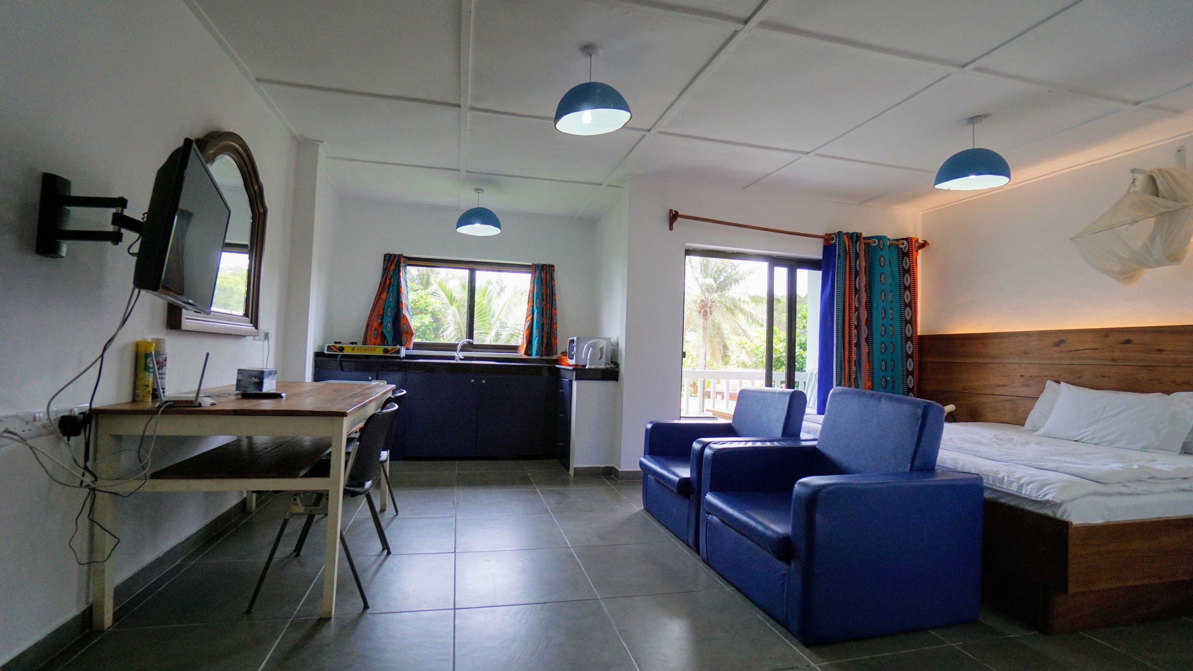 A room with a bed, a desk, and a chair. There is a window with curtains, a television mounted on the wall, and a ceiling light.