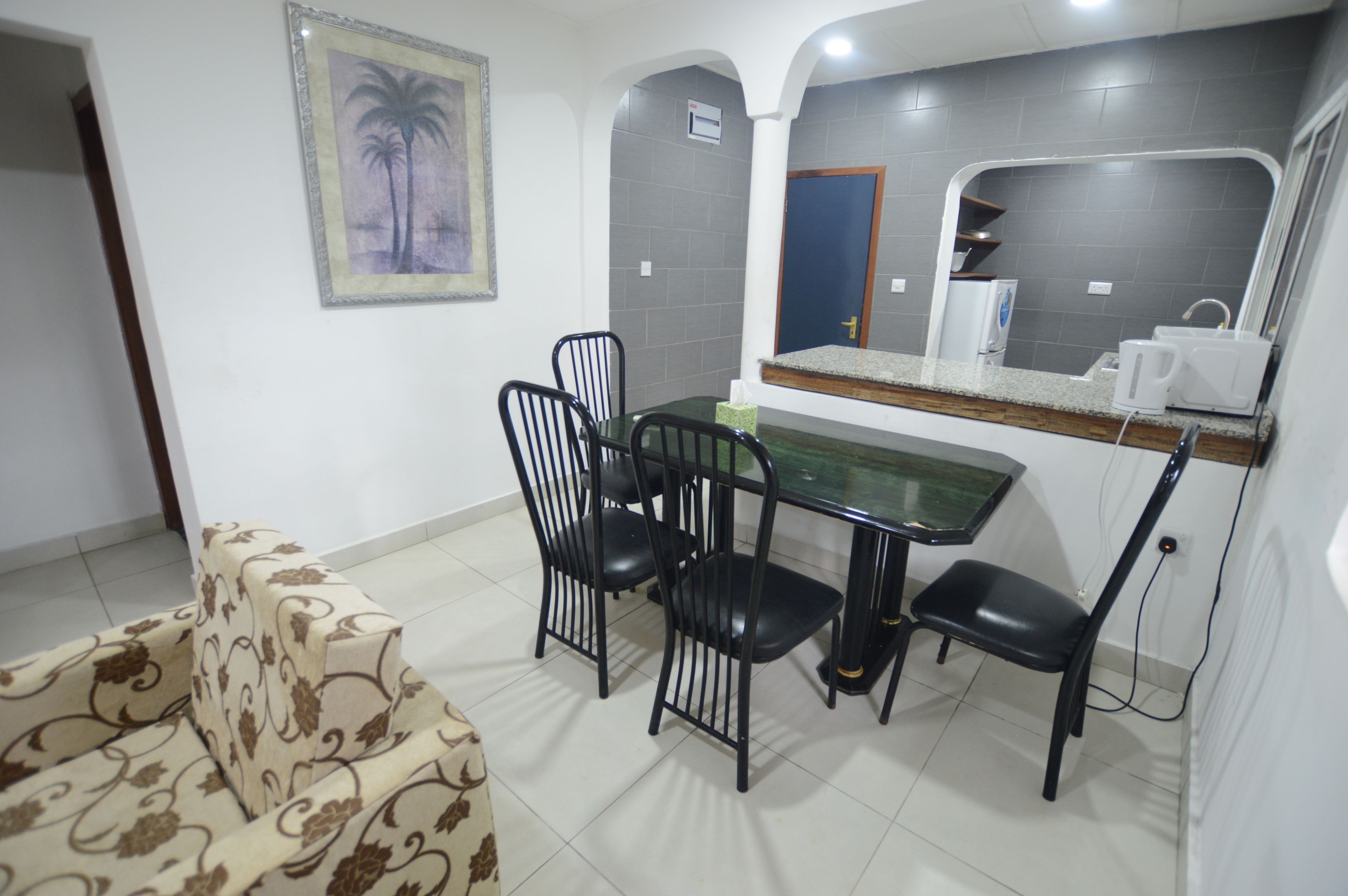 A dining area with a table and four chairs, a sofa, a framed picture on the wall, and a counter with a mirror and a coffee maker.