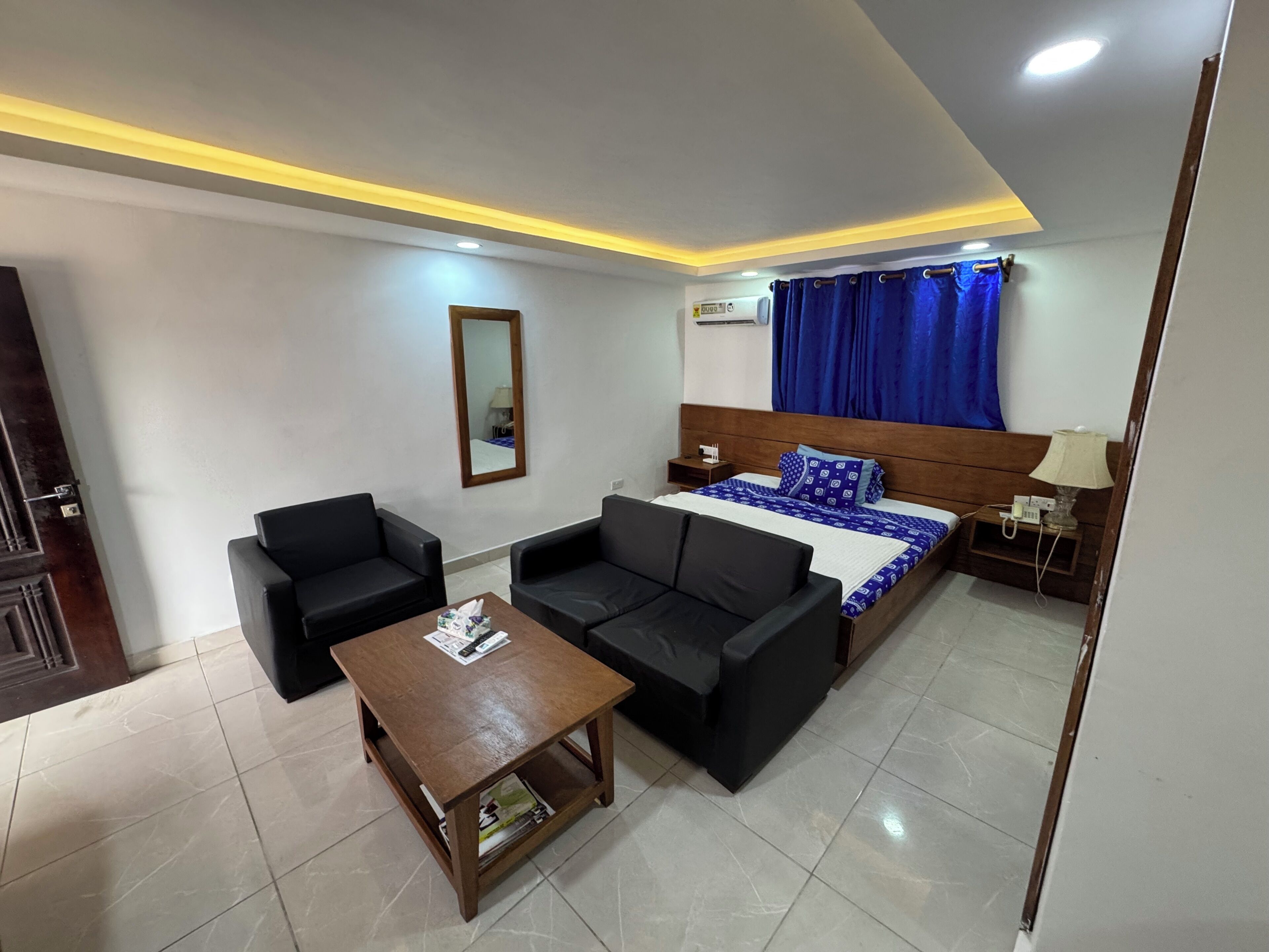 A hotel room with a bed, two black sofas, a wooden coffee table, a mirror, a nightstand, and a lamp.