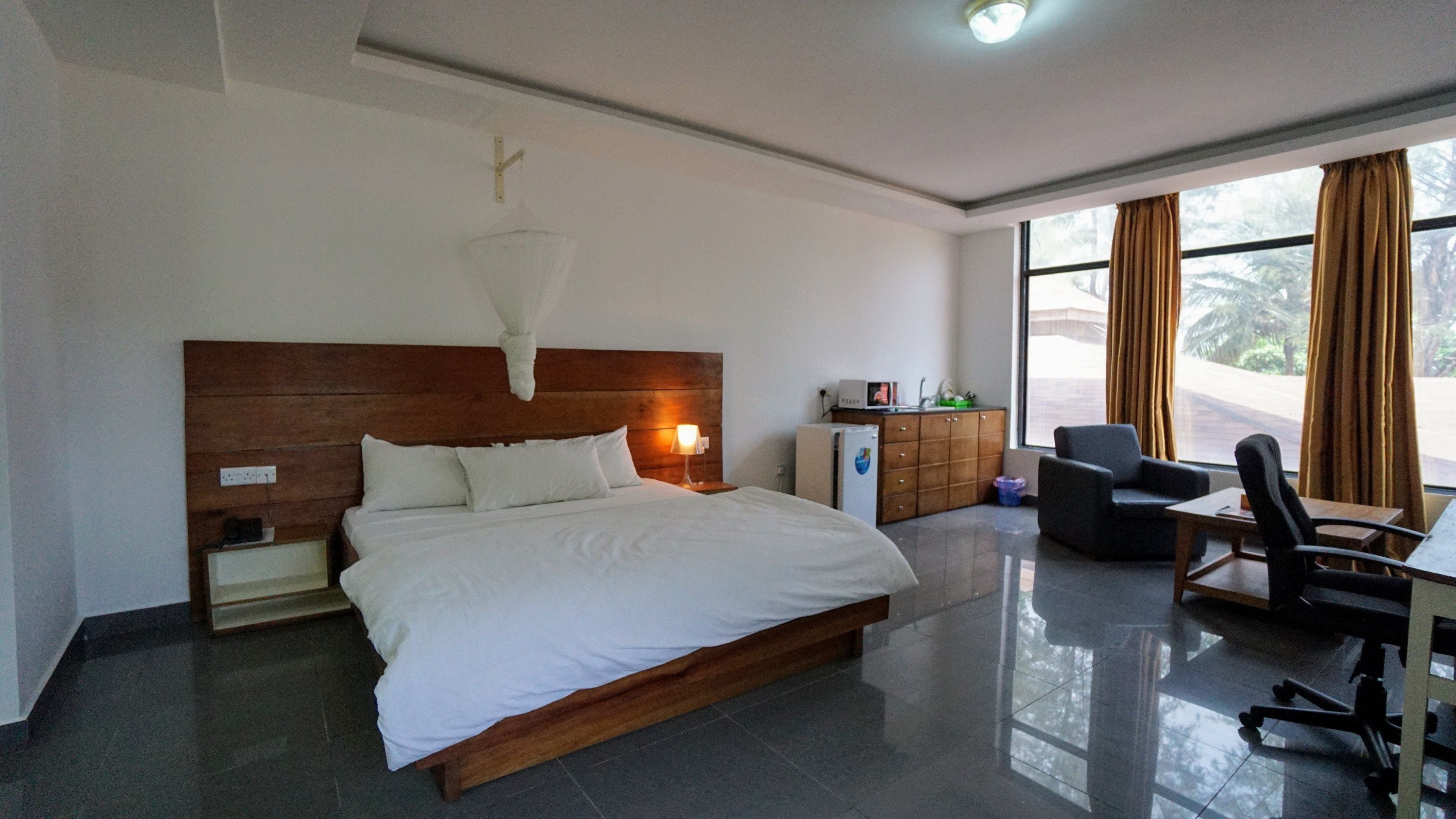 Una habitación de hotel moderna con una cama grande, un cabecero de madera, un escritorio con una silla, un refrigerador pequeño y una ventana con cortinas.