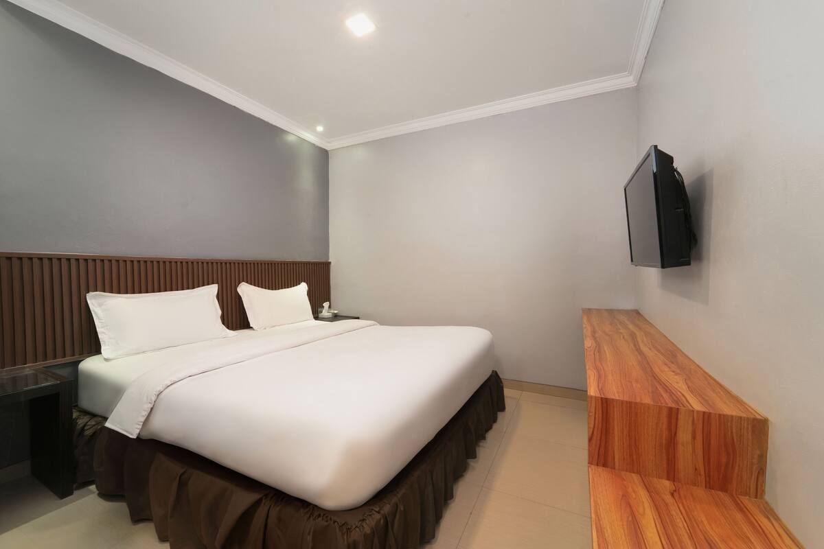 A hotel room with a large bed, a wooden nightstand, a TV mounted on the wall, and a wooden bench.