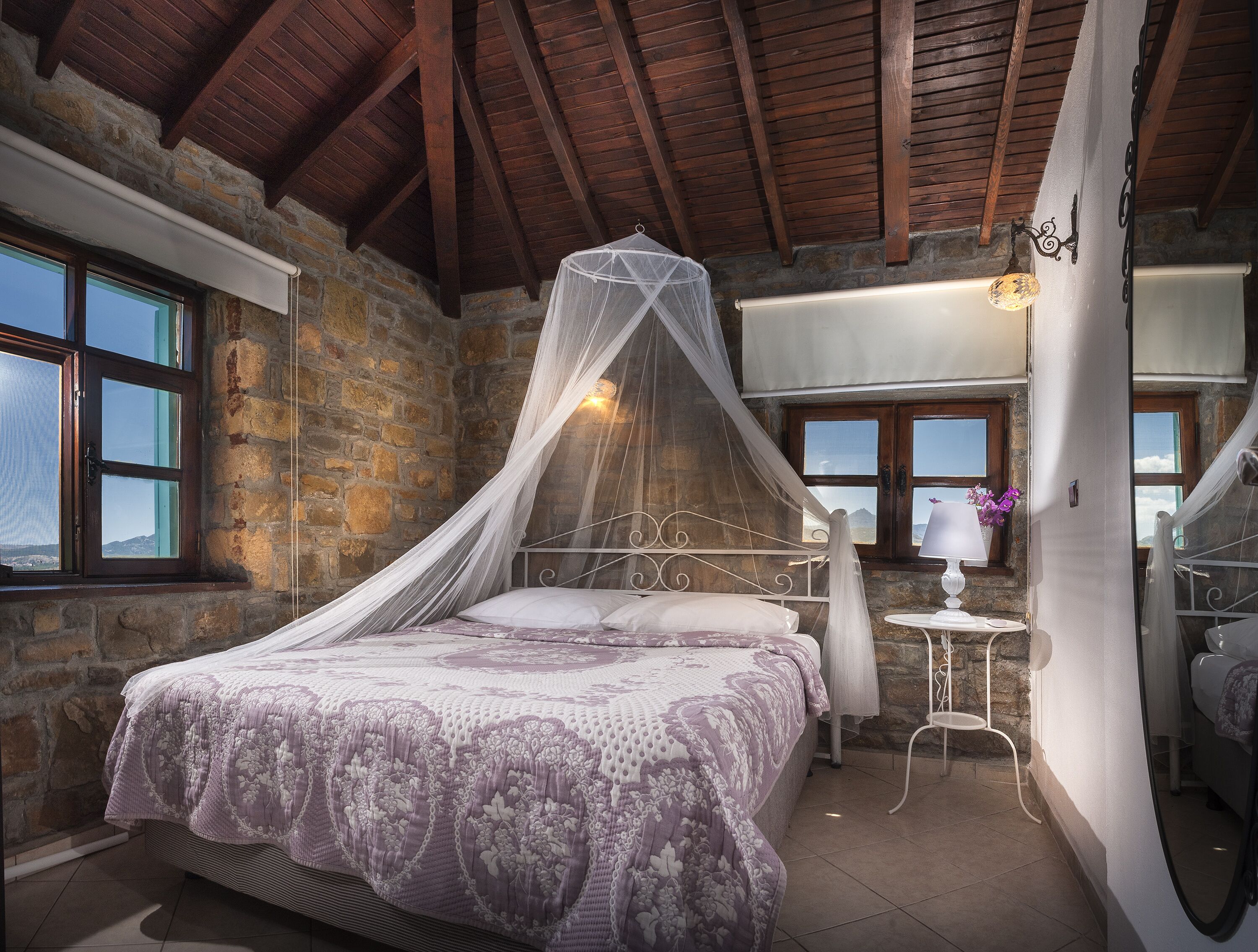 A bedroom with a stone wall, a bed with a canopy, a small table with a lamp, and a view through a window.