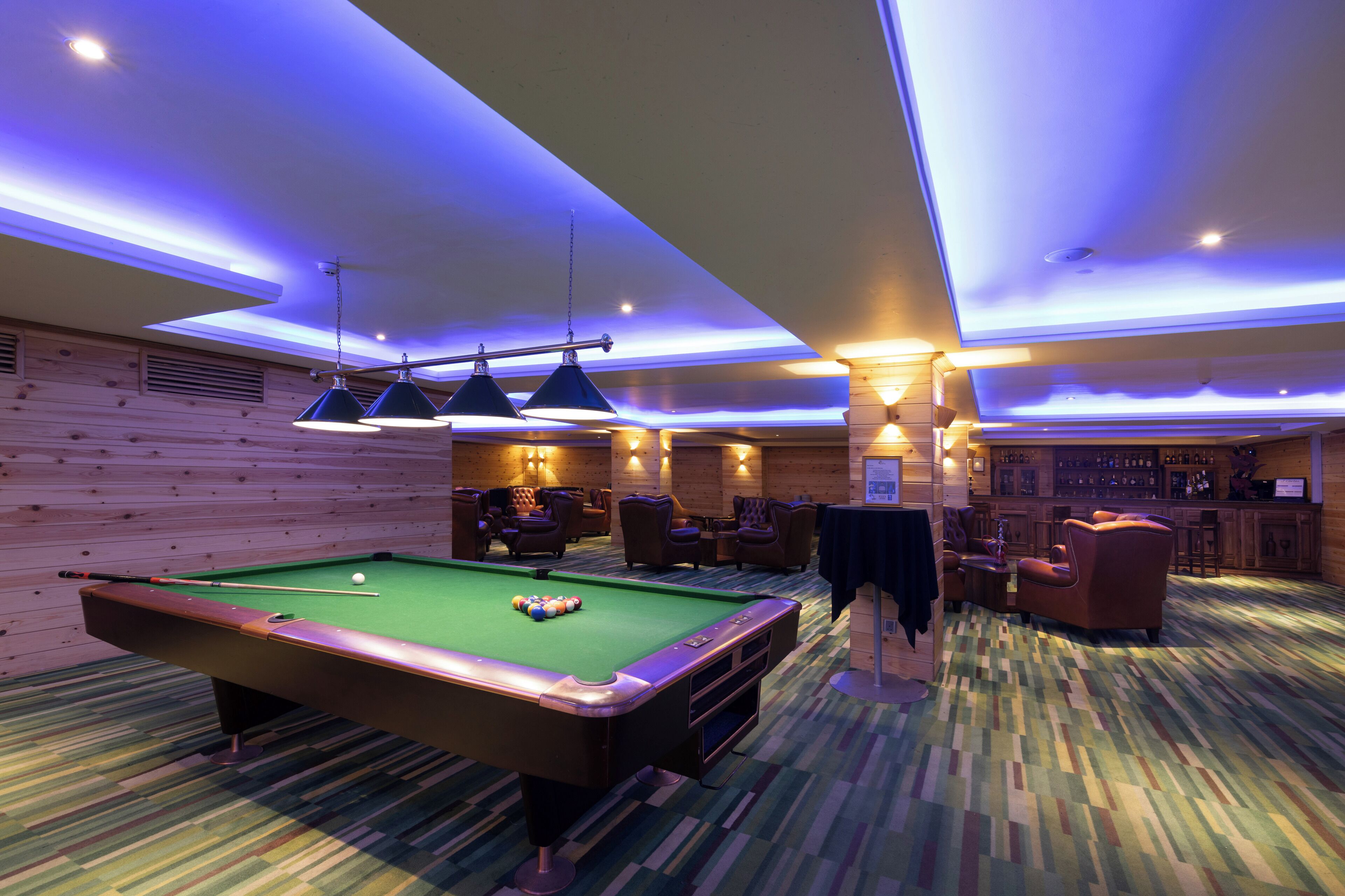 A pool table in a room with wooden walls, a bar area, and seating.