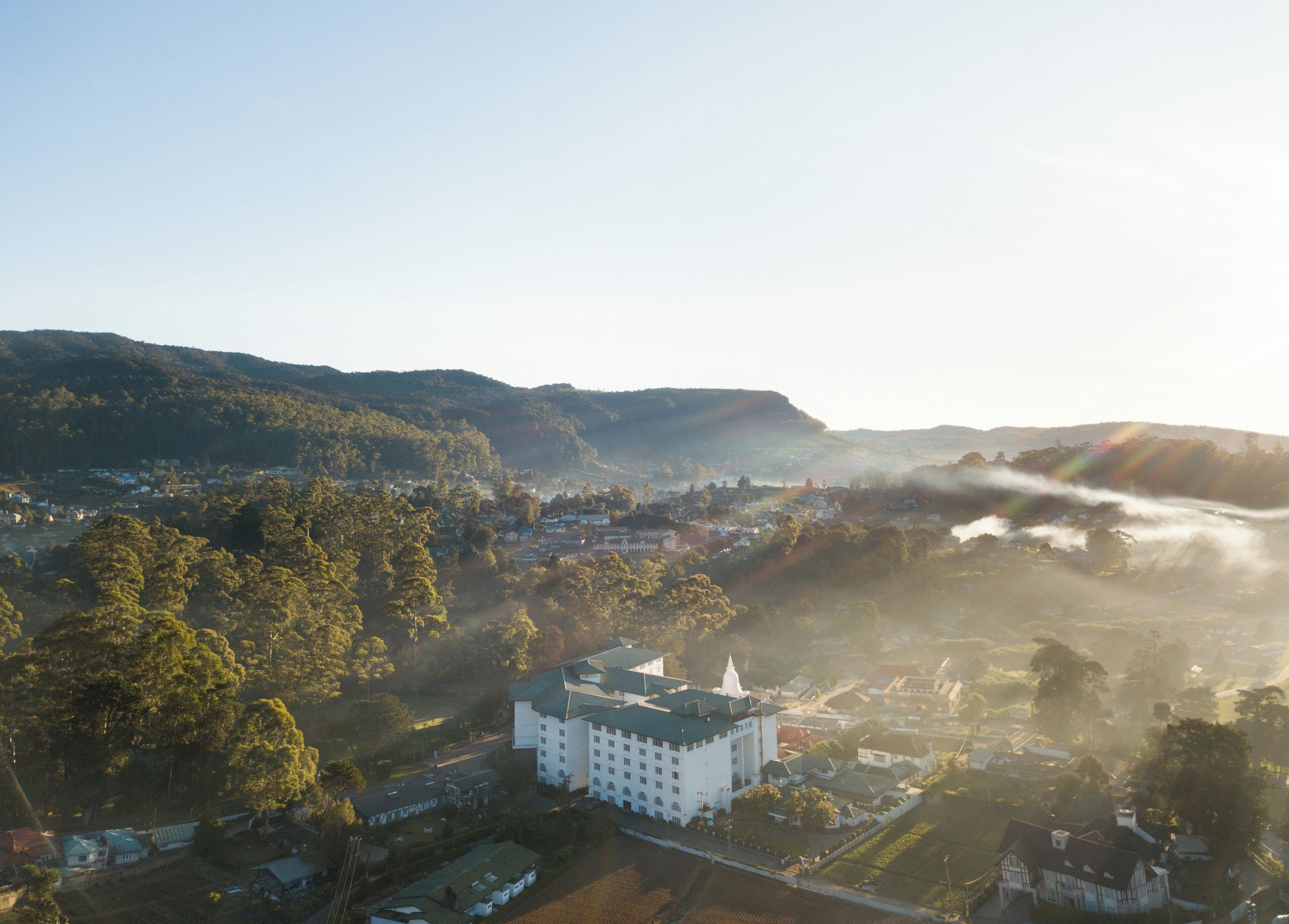An aerial view of a town with a prominent white building with a green roof, surrounded by lush greenery and hills in the background.