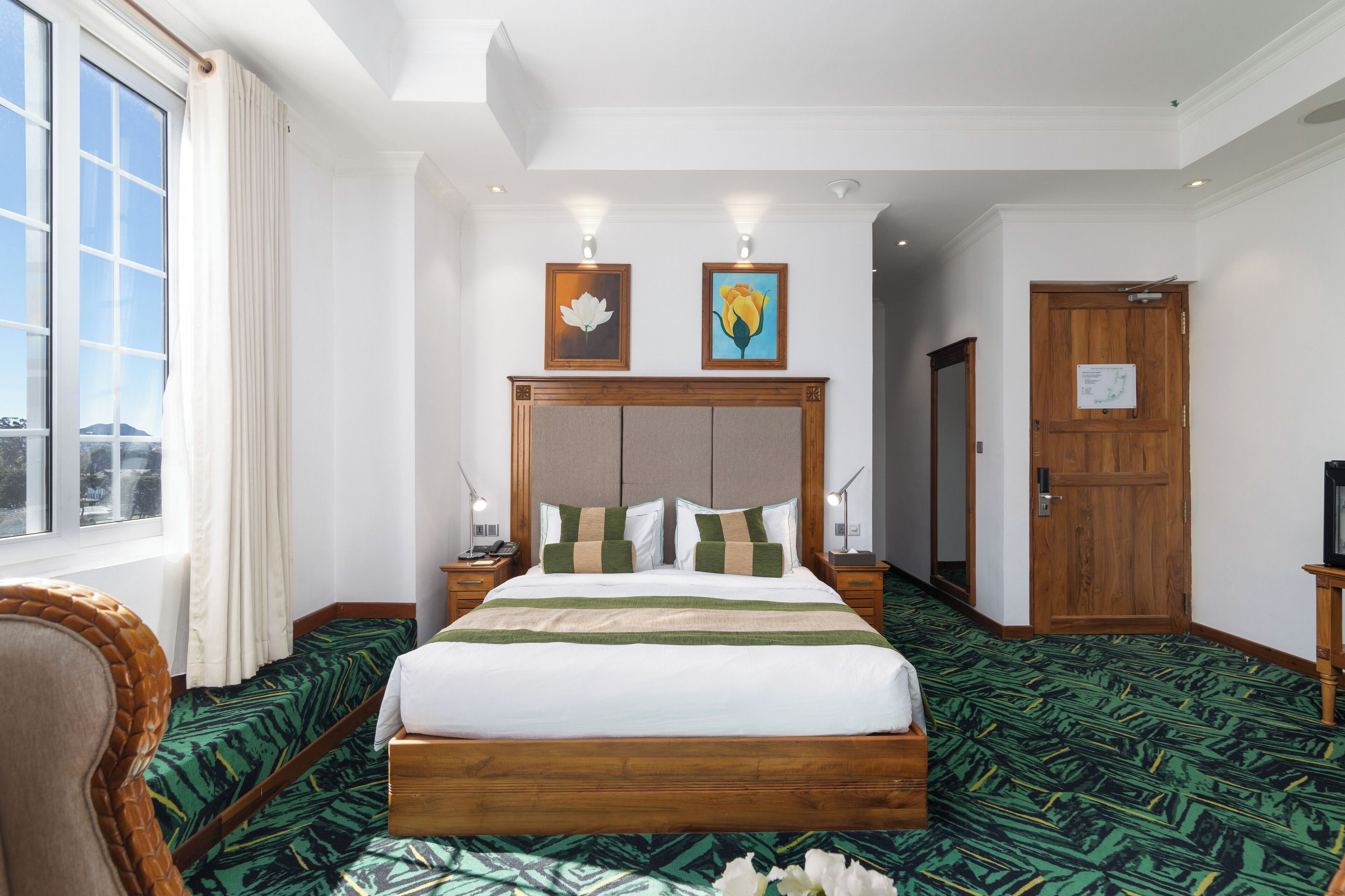 Une chambre d’hôtel avec un grand lit, une tête de lit en bois, deux tables de chevet avec des lampes, une fenêtre avec des rideaux et une porte.