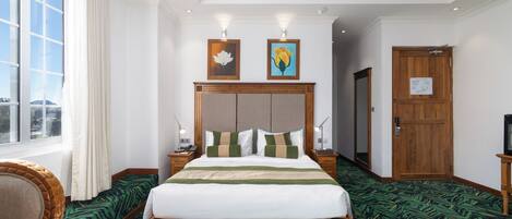 Una habitación de hotel con una cama grande, un cabecero de madera, dos mesitas de noche con lámparas, una ventana con cortinas y una puerta.