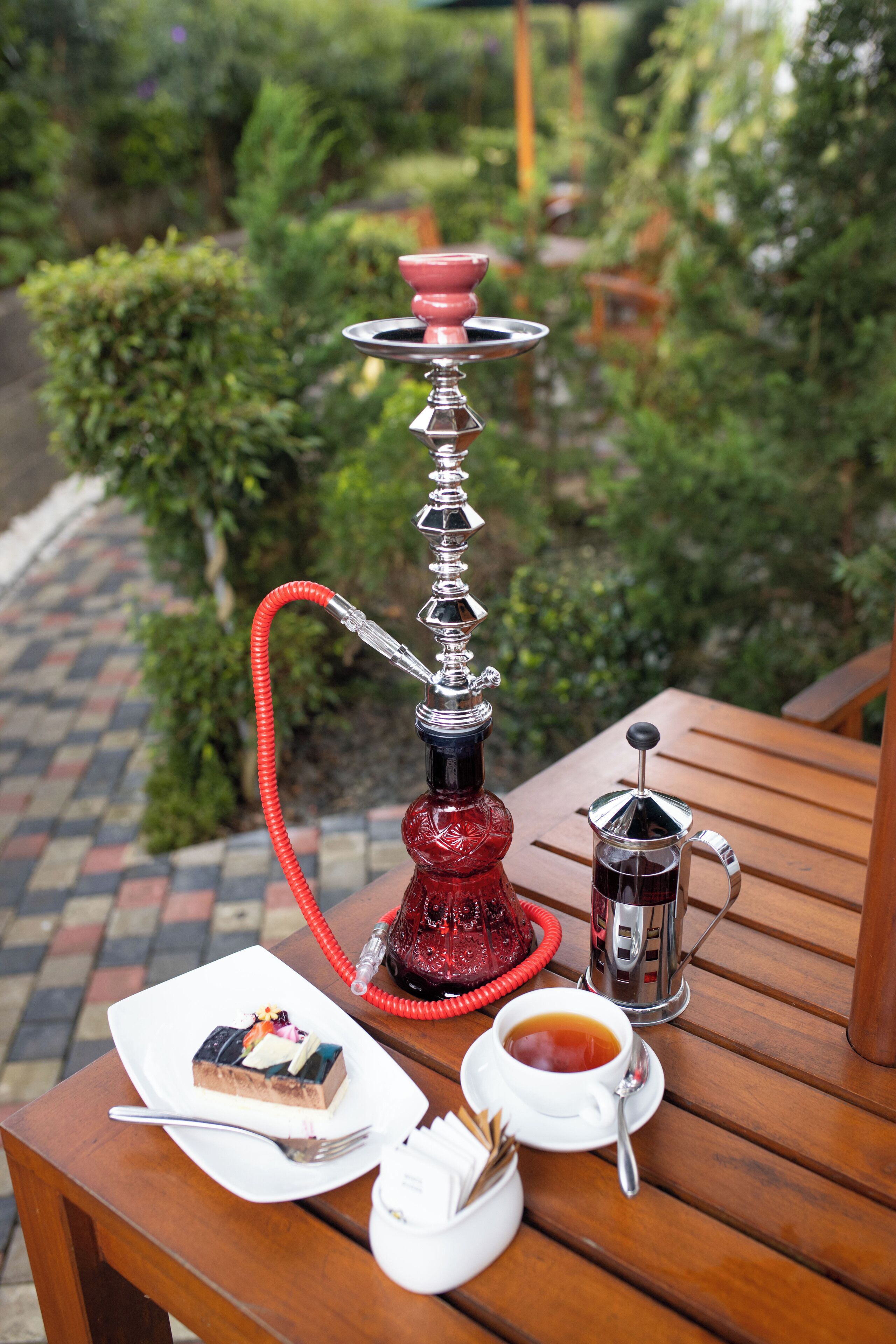 A hookah on a wooden table with a plate of food, a cup of tea, and a small container of sugar.