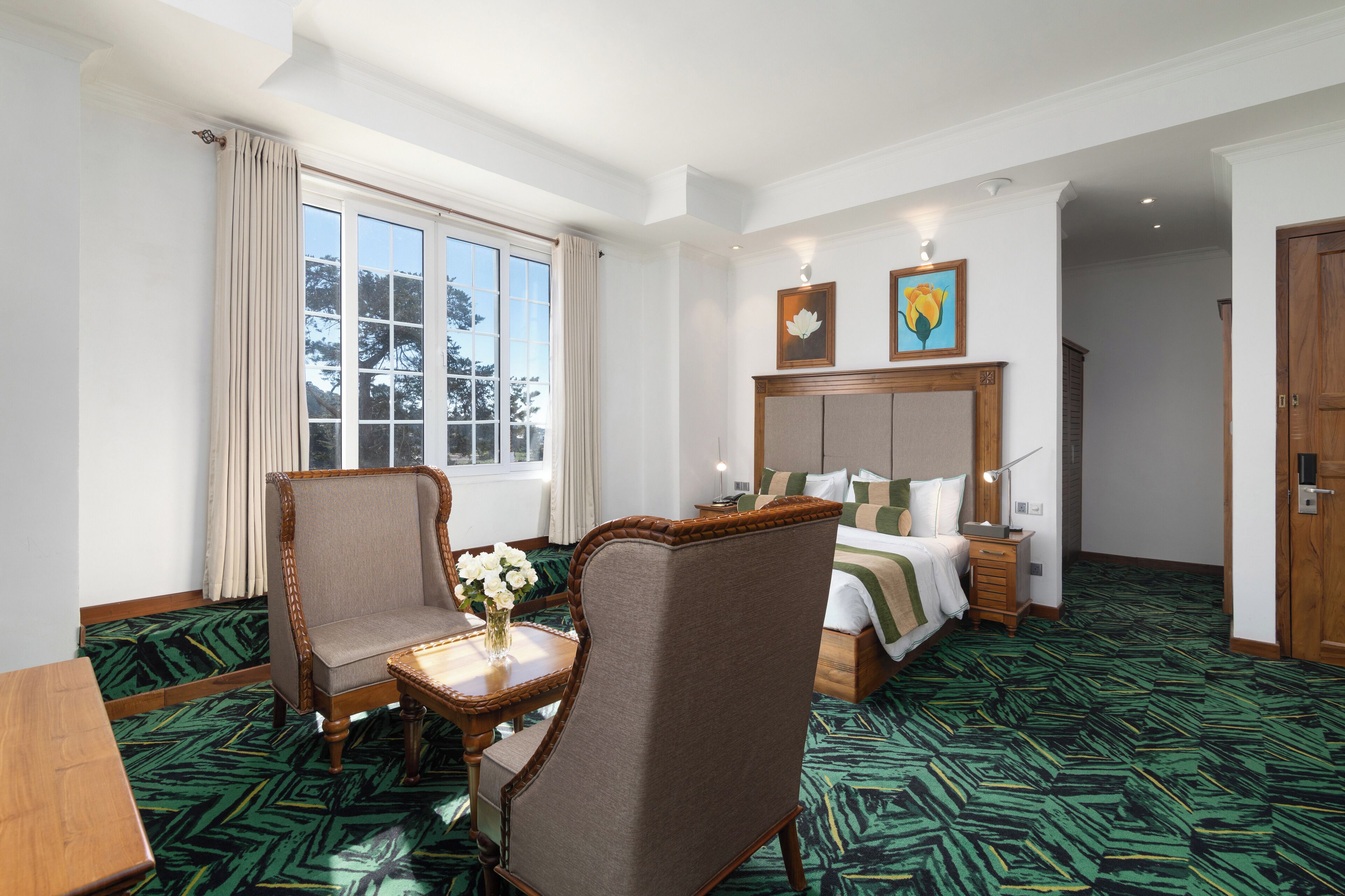 A hotel room with a large bed, a desk with a chair, a window with curtains, and a green patterned carpet.