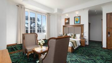 A hotel room with a large bed, a desk with a chair, a window with curtains, and a green patterned carpet.