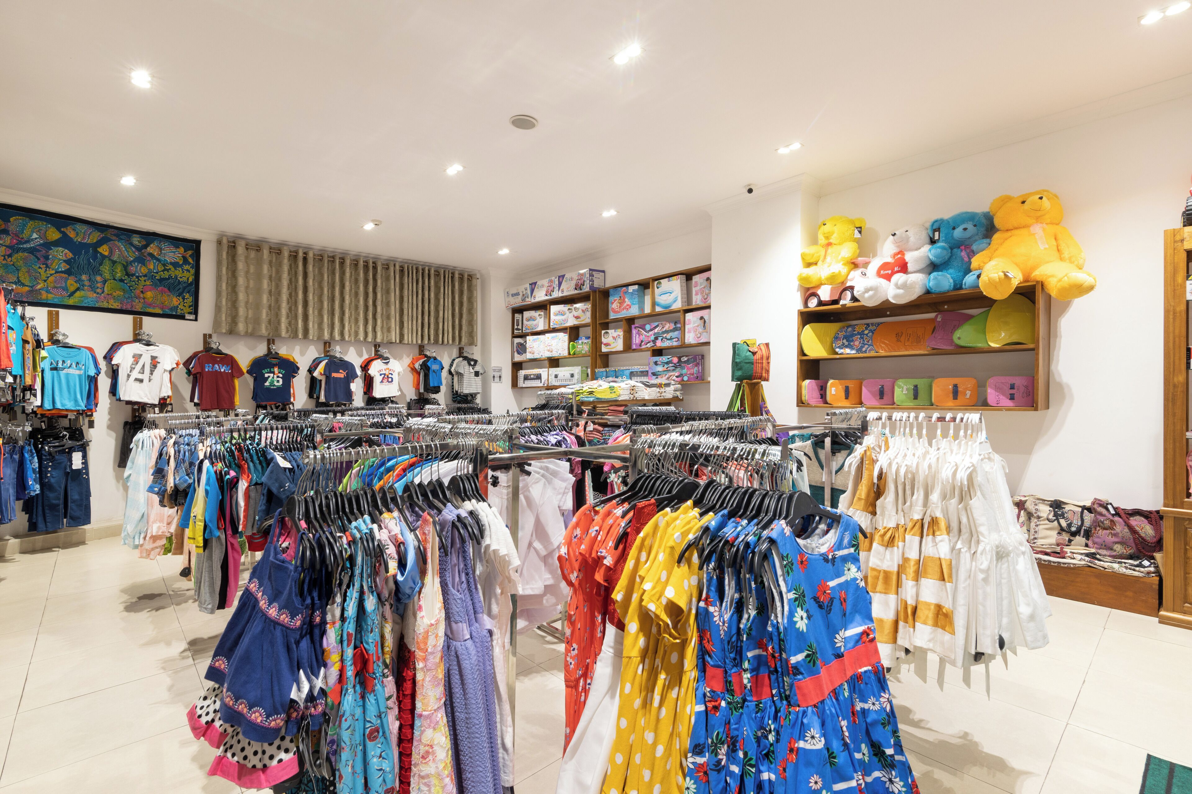 A clothing store with racks of dresses and shirts, shelves with stuffed toys, and a large painting on the wall.
