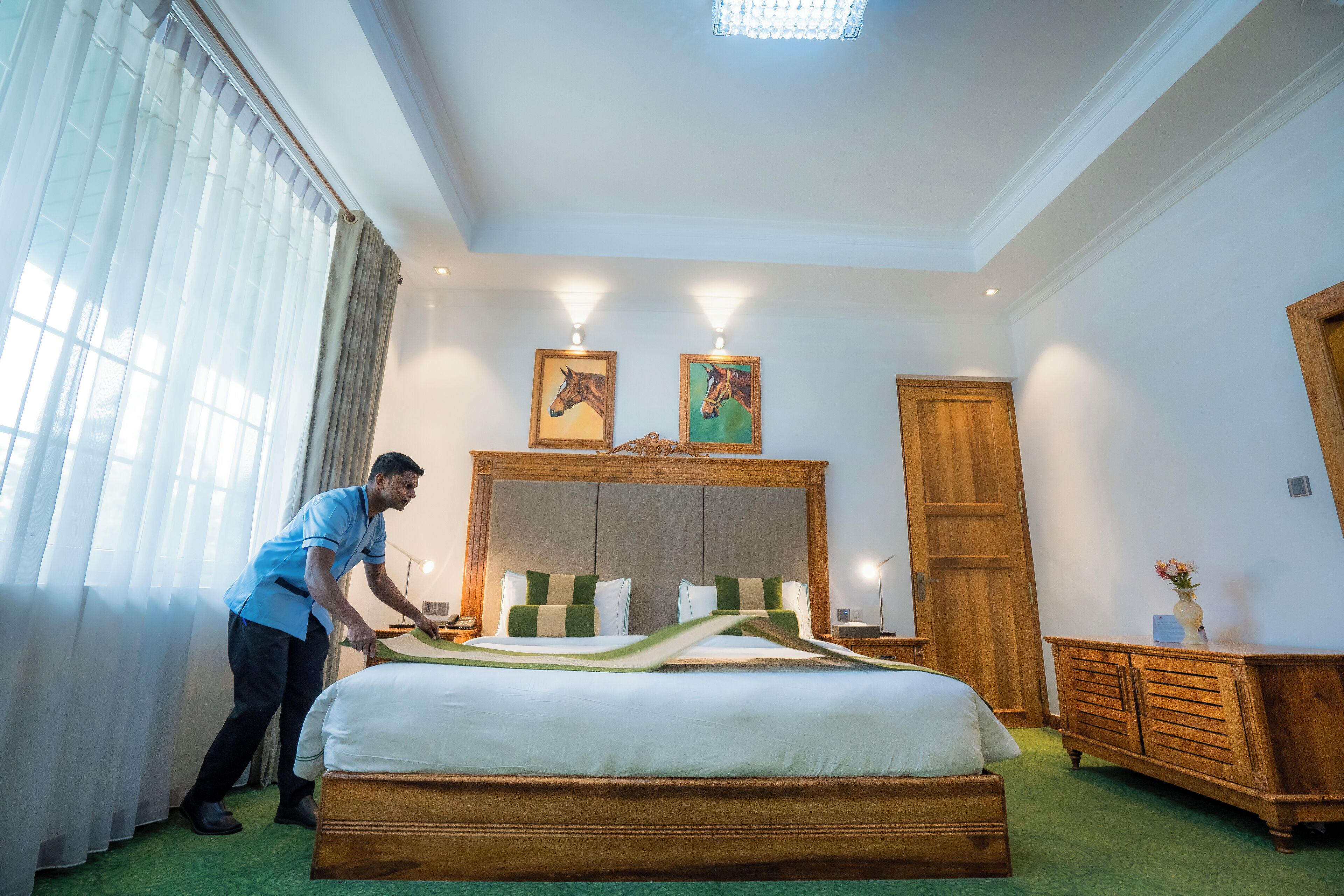 A hotel room with a large bed, wooden headboard, two framed horse paintings, a wooden wardrobe, and a green carpet.