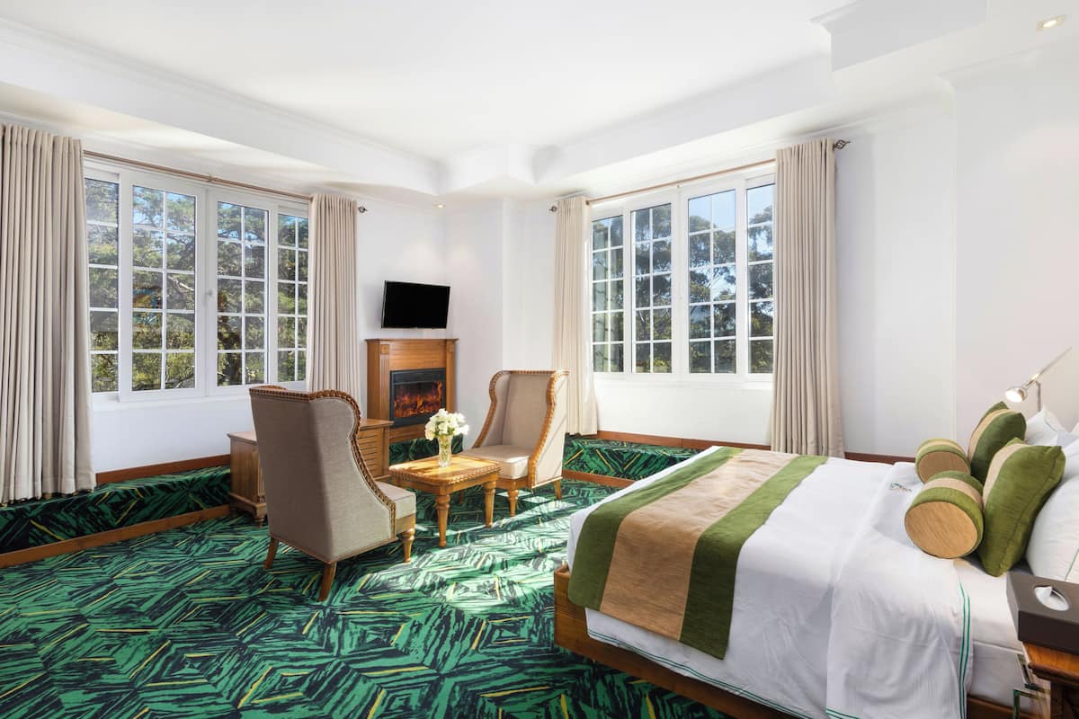 Une chambre d’hôtel avec un grand lit, deux fauteuils, une petite table avec un vase, une cheminée et de grandes fenêtres avec des rideaux.