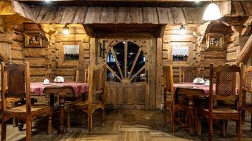 A rustic wooden dining room with a central wooden door, flanked by two windows with lace curtains.