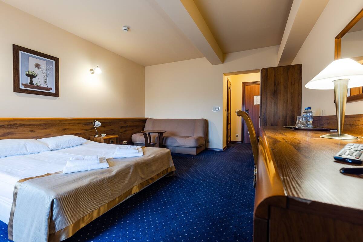Ett hotellrum med en säng, ett skrivbord med en lampa, en stol, en soffa och en inramad tavla på väggen.