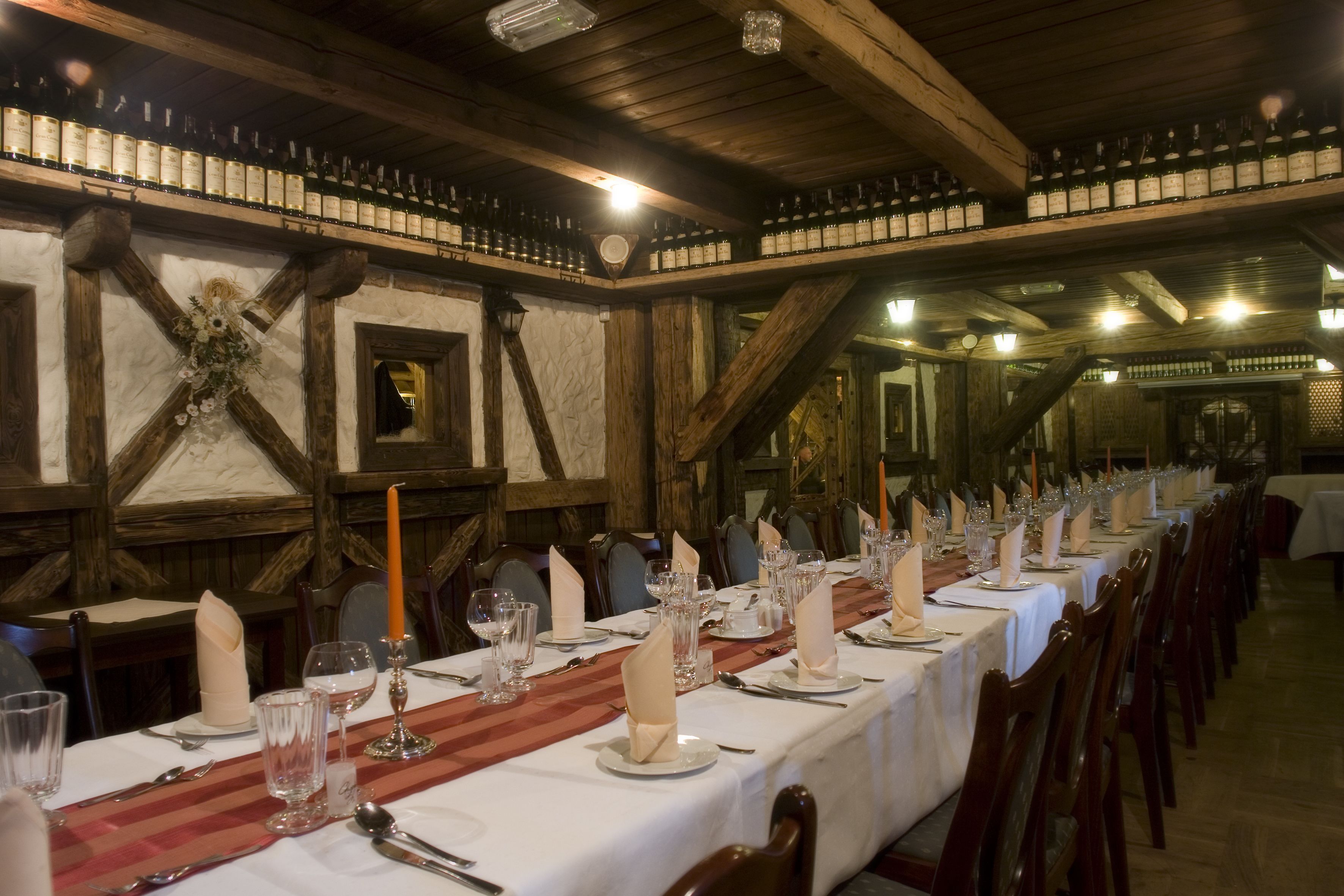 Una larga mesa de comedor dispuesta con manteles blancos, velas y cristalería en un rústico salón de madera con vigas a la vista y un techo alto.