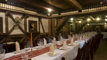 Una larga mesa de comedor dispuesta con manteles blancos, velas y cristalería en un rústico salón de madera con vigas a la vista y un techo alto.