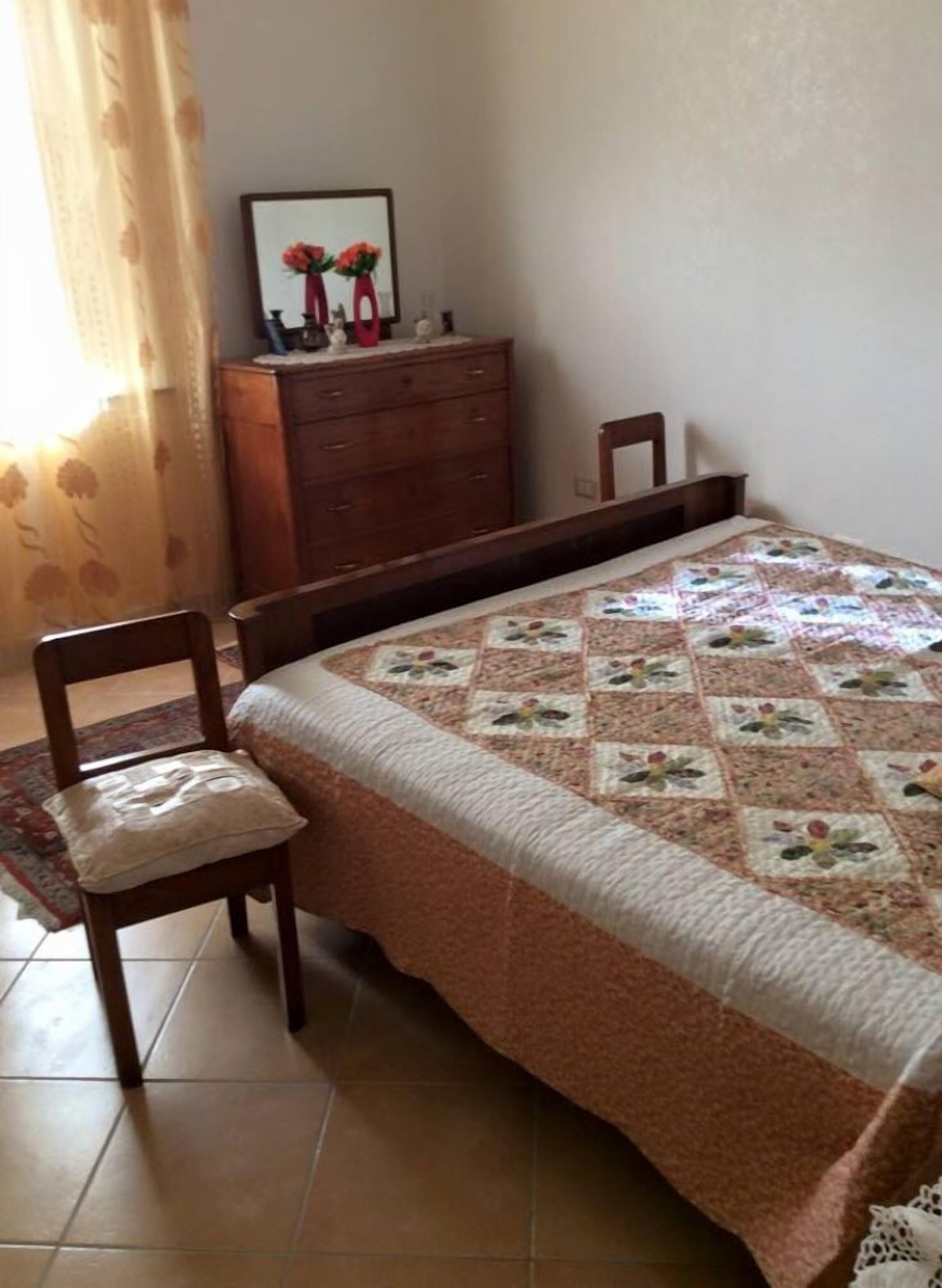 A bedroom with a bed, a wooden dresser, a chair, and a framed picture on the dresser.