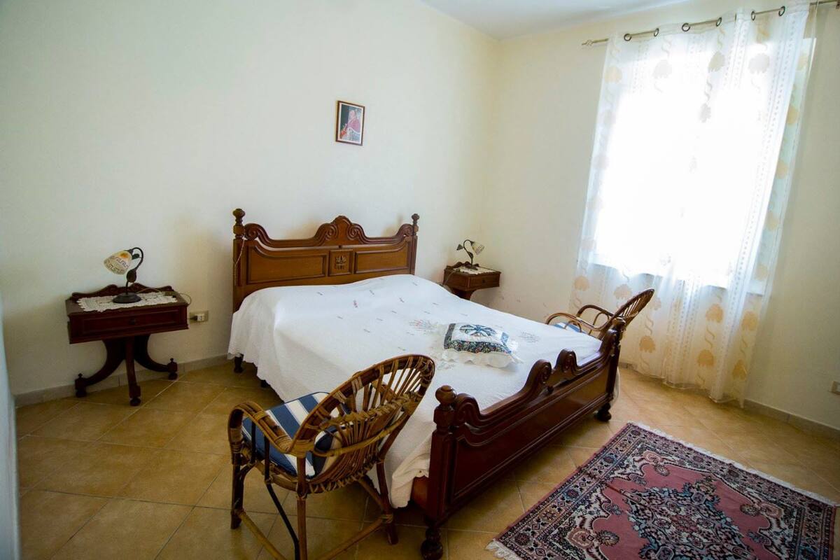 Een slaapkamer met een houten bed, een nachtkastje, een stoel, een vloerkleed en een raam met gordijnen.