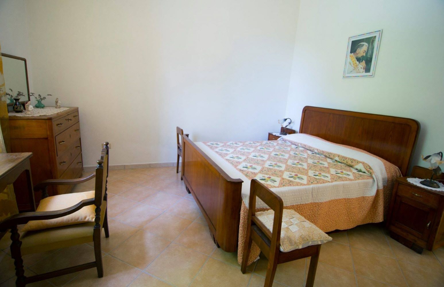 Um quarto com uma cama de madeira, uma cadeira de madeira, uma mesinha de cabeceira e um aparador.